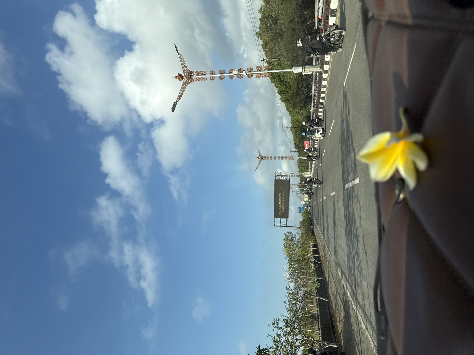 The image depicts a bright, daytime scene on a highway in Unknown, Indonesia, with a clear blue sky dotted with white, fluffy clouds. In the foreground, a yellow flower rests on a dark brown, textured surface, possibly a car dashboard. The highway stretches into the distance, with multiple lanes of traffic, primarily consisting of motorcycles. Several individuals are seen riding motorcycles, some wearing helmets and backpacks. The road is lined with trees and vegetation, and ornate lampposts with decorative tops are visible along the side. A large digital sign with text in Indonesian is positioned above the highway. The perspective suggests the photo was taken from inside a moving vehicle.