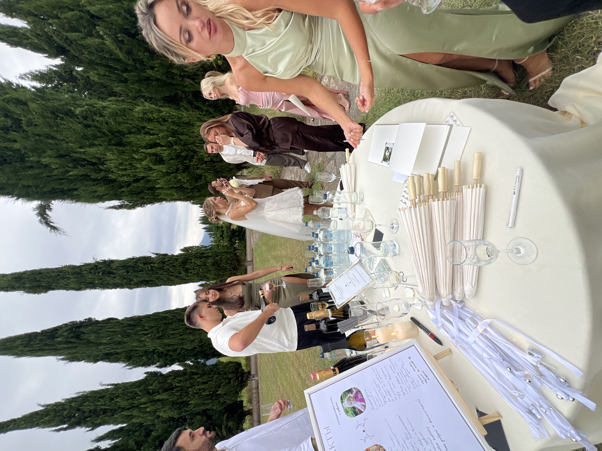 The image depicts an outdoor wedding reception in Napareuli, Georgia, with a table laden with champagne bottles, water bottles, glasses, and small white parasols. Several guests are gathered around the table, some engaged in conversation and others observing the proceedings. A man in a white polo shirt is pouring champagne into a glass, while a woman in a green dress stands next to him. A bride in a white dress and a groom in a brown suit are also visible in the background. The setting appears to be a garden with tall cypress trees and a cloudy sky, suggesting a potentially cool day.