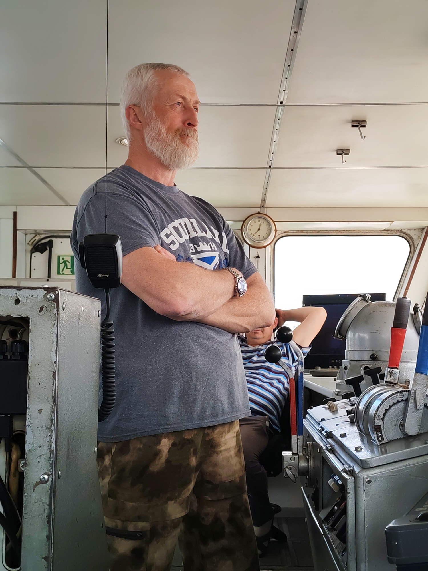 The image depicts the interior of a ship's bridge or control room, likely in Lumbushi, Russia. In the foreground, a mature man with a prominent white beard and grey hair stands, his arms crossed, looking out of a large, bright window to the right. He wears a grey t-shirt, part of which displays "SCOTLAND" text, and camouflage-patterned trousers, with a watch visible on his left wrist. His expression appears calm and observant.
To the left of the standing man, a black microphone with the word "Marry" printed on it hangs from a coiled cord, attached to a gray, industrial-looking console. In the background, behind the standing man and to his right, another person wearing a striped shirt is partially visible, seated at a complex control panel featuring various levers with red and blue handles. Above the window, a round analog clock indicates the time. The ceiling is paneled in white with visible metal tracks and small hooks. On the wall to the far left, an emergency exit sign with the running man symbol is visible. The bright light from the window suggests it is daytime, with potentially clear weather outside.