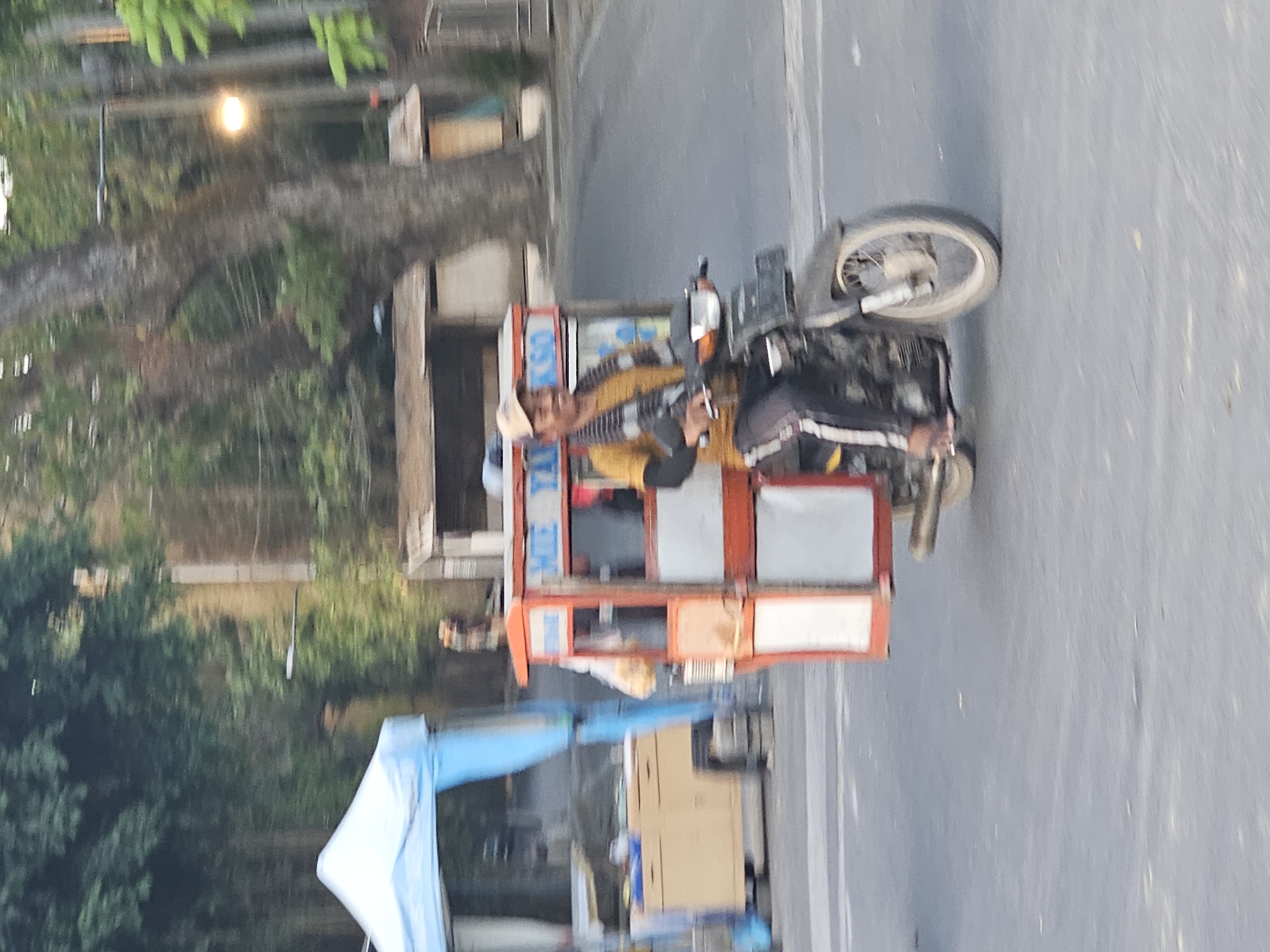 A man is riding a motorcycle with a food cart attached to the rear in Denpasar, Indonesia. The food cart is red and white and has "MIE YAKSO" written on it. The man is wearing a yellow shirt, dark pants, and a beige cap. He is smiling and looking forward. The motorcycle is black and has a license plate. The scene appears to be a street with trees and buildings in the background. The lighting suggests it is daytime.