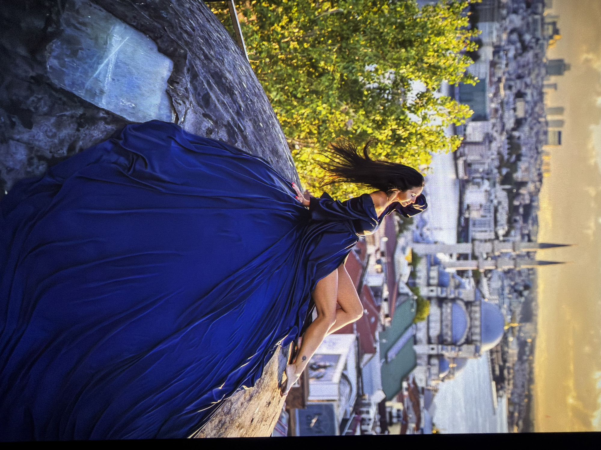 A woman in a flowing dark blue dress is posed on a large, rough-textured rock formation. Her body is angled, with one leg extended and her head tilted back. Her hair is long and dark, flowing around her shoulders. Behind her, a cityscape unfolds, featuring numerous buildings and a prominent mosque with domes and minarets. The sky is illuminated with the warm glow of sunset, casting a golden hue over the scene. Green trees are visible to the left of the woman. The image appears to be a posed photograph. The location is not Kadıköy, Turkey, but rather Istanbul, Turkey, likely near the Blue Mosque or a similar prominent landmark.