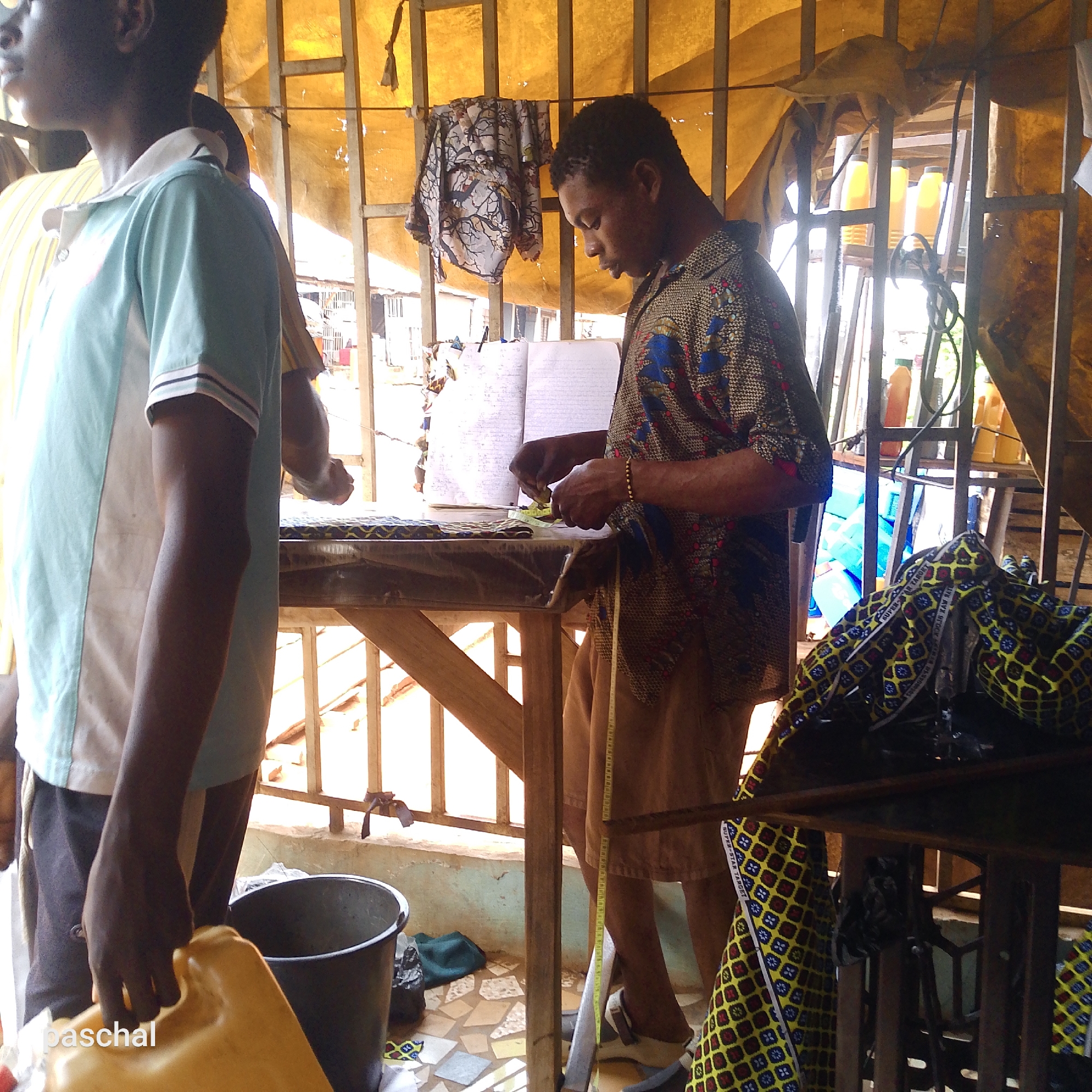 The image appears to depict a tailor's workshop, possibly in Nsugbe, Nigeria, given the prompt.
Two men are present. One man, seen in profile on the left, wears a light blue and white shirt and dark shorts. He carries a yellow jerrycan. The other man stands at a wooden table, wearing a patterned shirt and brown shorts. He is measuring fabric on the table with a measuring tape and consulting an open notebook.
The workshop is enclosed by what seems to be a metal grid and a translucent fabric awning. Behind the tailor, clothing is hung on the grid. Sewing supplies and fabrics are visible around the table, including a sewing machine on the right. A bucket is near the jerrycan. The floor is tiled. The overall impression is of a small, functional workspace.