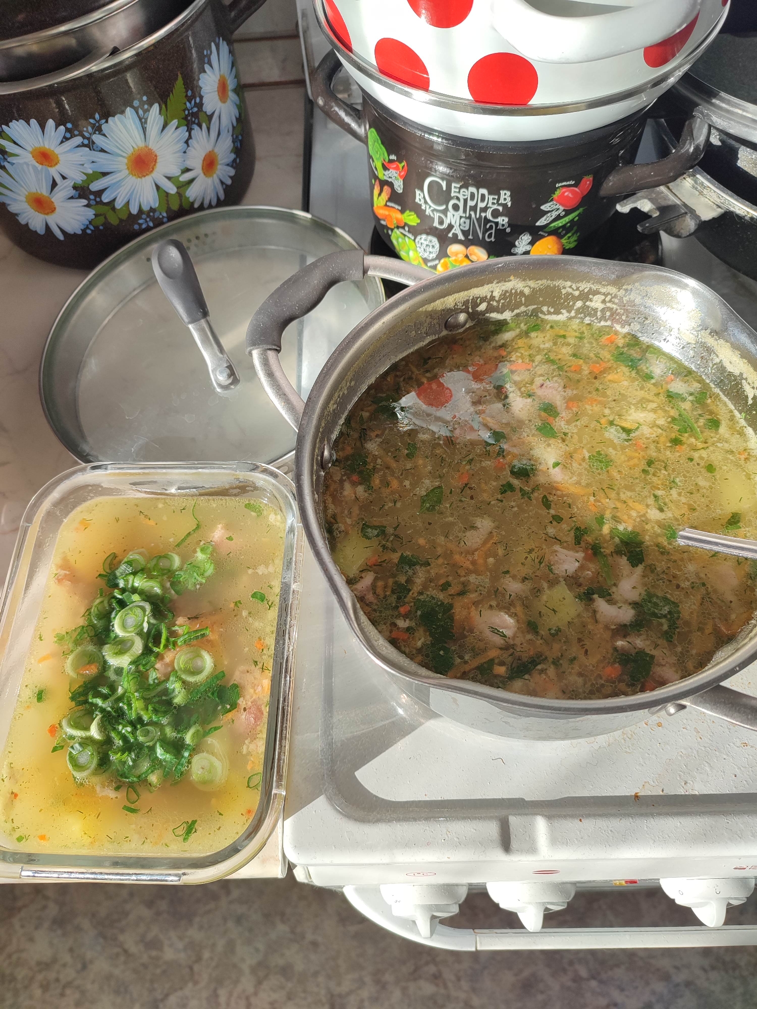 A high-angle shot depicts a domestic kitchen scene in Unknown, Unknown. Three pots are visible: one with a daisy pattern, another black with vegetable illustrations, and a third with red polka dots stacked on top. A stainless-steel saucepan filled with soup containing greens and vegetables rests on a white stovetop. Next to it, a clear glass container holds another serving of soup, topped with fresh, chopped green onions. A stainless-steel lid with a gray handle sits nearby.