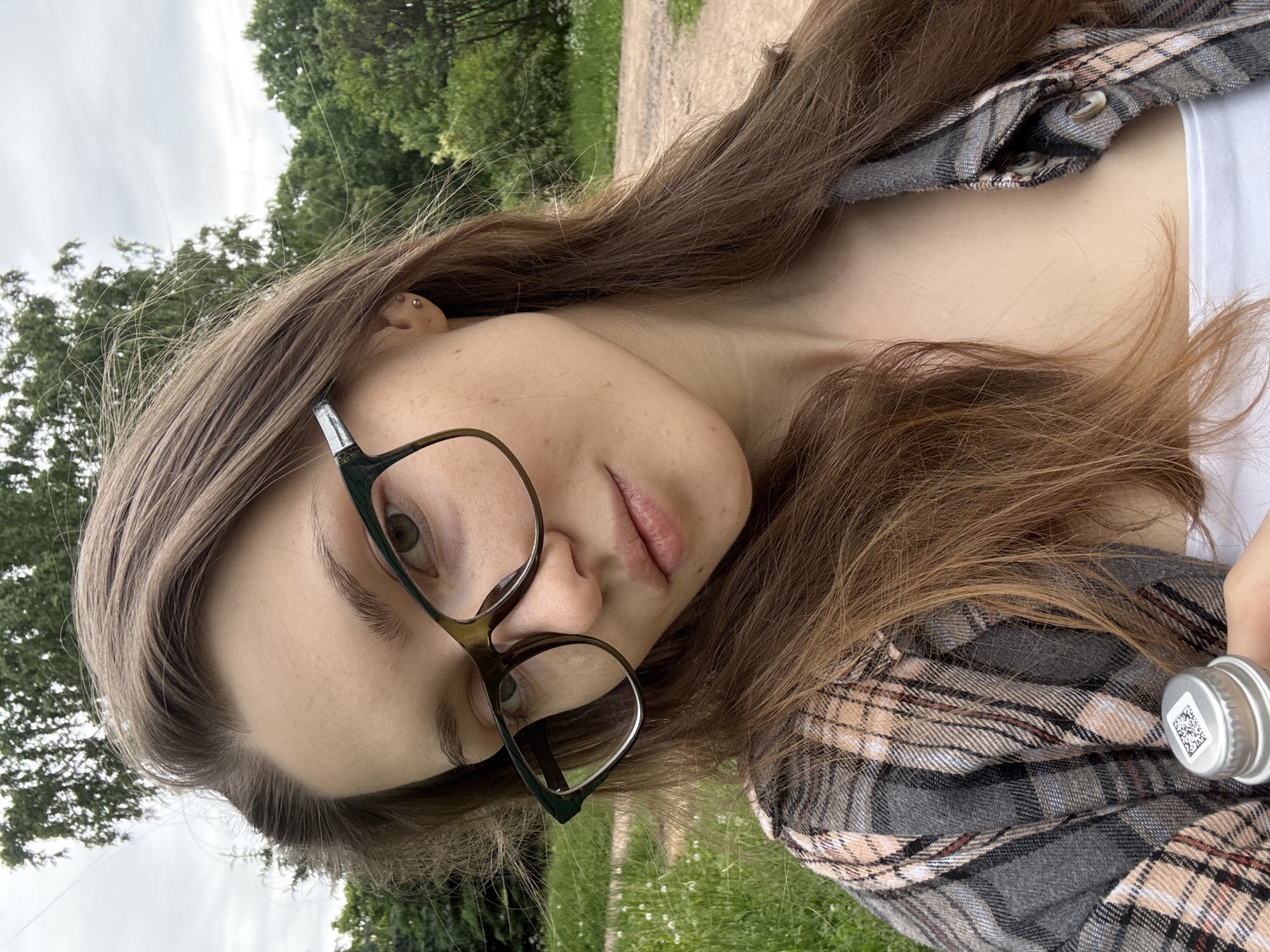 A young woman with long brown hair and glasses is taking a selfie outdoors. She is wearing a plaid shirt over a white top. In her hand, she is holding a bottle cap with a QR code. The background shows trees and a dirt path, suggesting a park or natural area. The lighting is overcast.