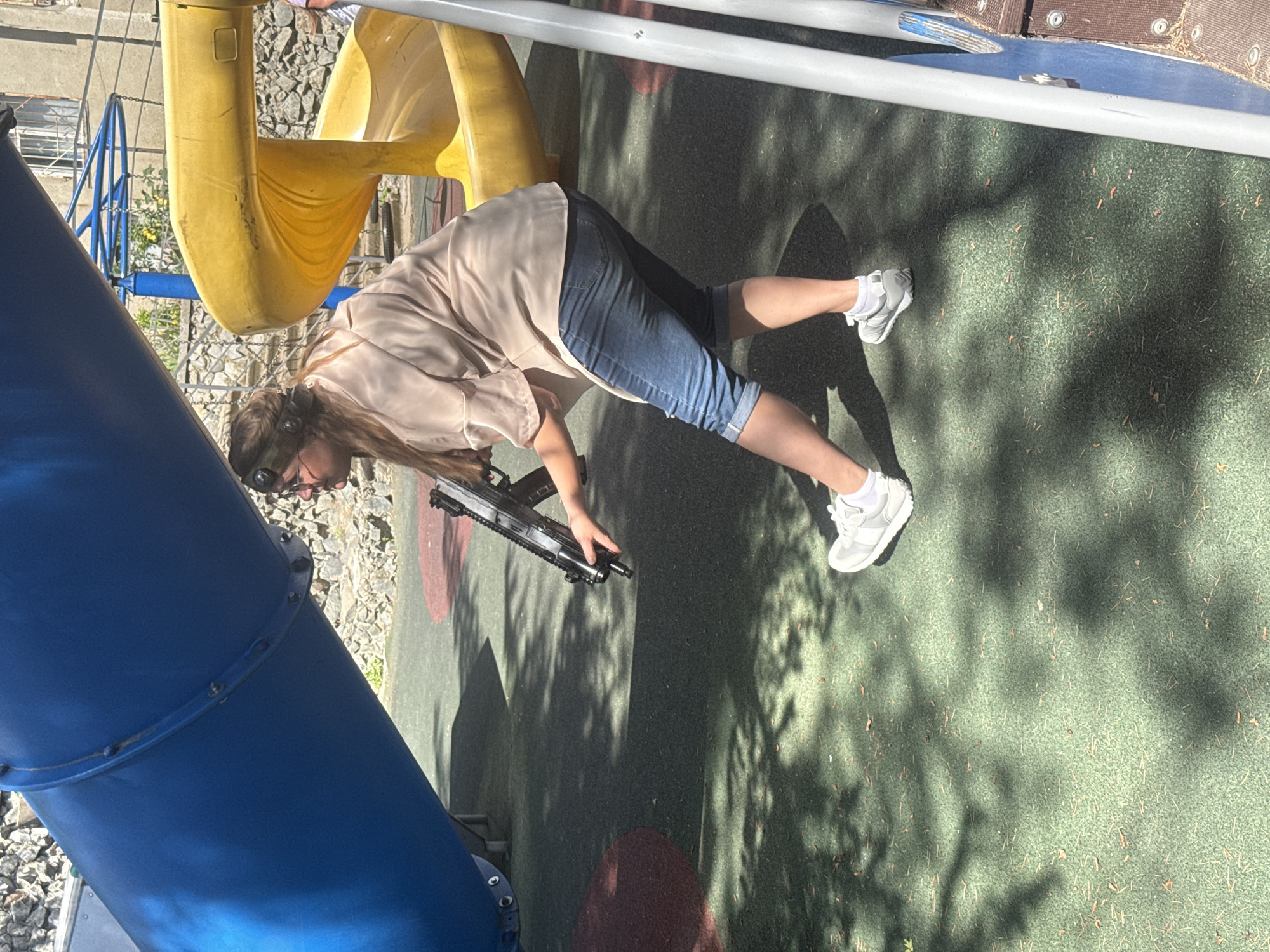 A person stands on a playground, dressed in a tan shirt and denim shorts. They are holding a toy gun, positioned in front of a blue slide and a yellow play structure. The ground covering of the playground is green, with some red spots. The scene is shadowed by trees.