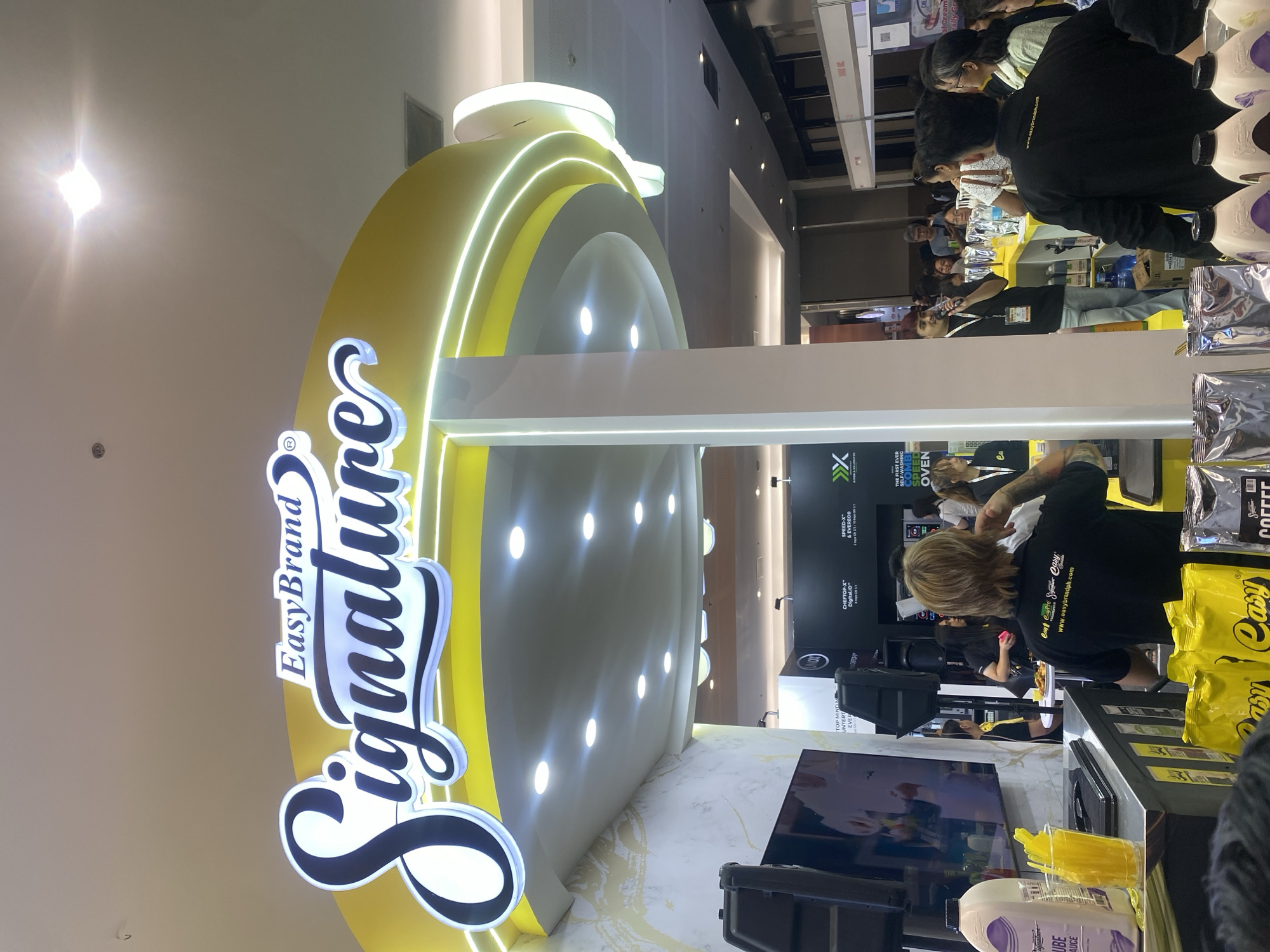 The image depicts an indoor event, likely a product launch or promotional booth, possibly at the SMX Convention Center in Davao City, Philippines. A large, illuminated sign prominently displays "EasyBrand Signature" in a stylized font, mounted on a curved yellow structure. Beneath this sign, a white pillar supports the overhead structure.
In the foreground, a counter displays various products, including a large bottle of "Signature Cube Sauce," cups with yellow stirrers, and bags of coffee. A person wearing a black t-shirt with the "EasyBrand" logo and website printed on the back is standing behind the counter, facing away from the camera. Other individuals are visible in the background, some interacting with the counter staff and others observing.
To the left of the counter, a large screen displays an image. Behind the counter, a stage area is set up with a man holding a microphone and speaking to an audience. Various booths and displays are visible in the middle ground and background, suggesting a larger exhibition or trade show. The overall environment appears to be well-lit with artificial lighting, typical of an indoor event space.