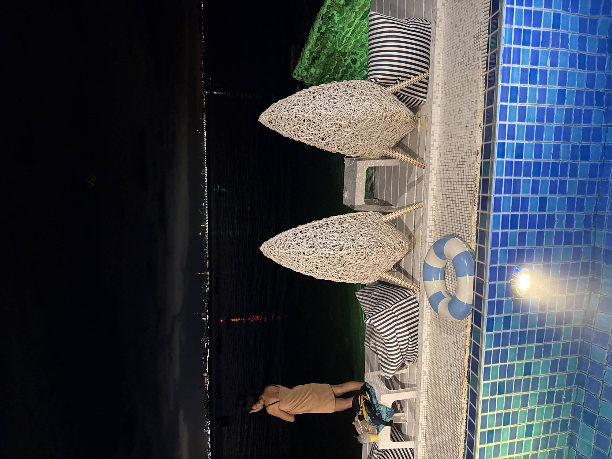 At night in Pattaya City, Thailand, a person in a short, tan dress lies horizontally on a gray-tiled deck next to a blue-tiled pool. They are near two white-and-blue striped pillows and a small white table holding a clear cup with a yellow liquid, a plastic bottle, and a blue bag. A blue-and-white striped life preserver hangs on the pool’s edge. The deck has two white-wicker lounge chairs, one topped with a green pillow. Across the dark water, city lights illuminate the horizon.