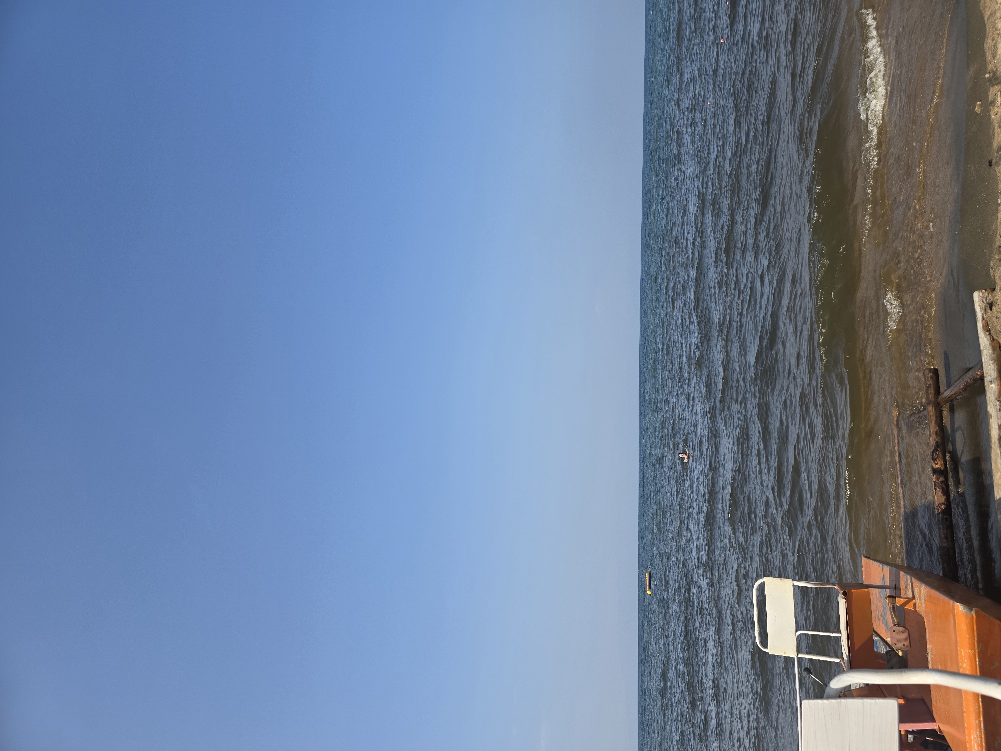 A wide shot captures a serene lake scene under a clear blue sky. In the foreground, an orange pedal boat with a white chair is partially visible on the sandy shore, its front end angled towards the water. The lake stretches out to the horizon, its surface rippled by a gentle breeze. Two distant figures are visible in the water; one is swimming, and the other is near a yellow float. The overall impression is one of a tranquil summer day at a lakeside in Maksimiha, Russia.