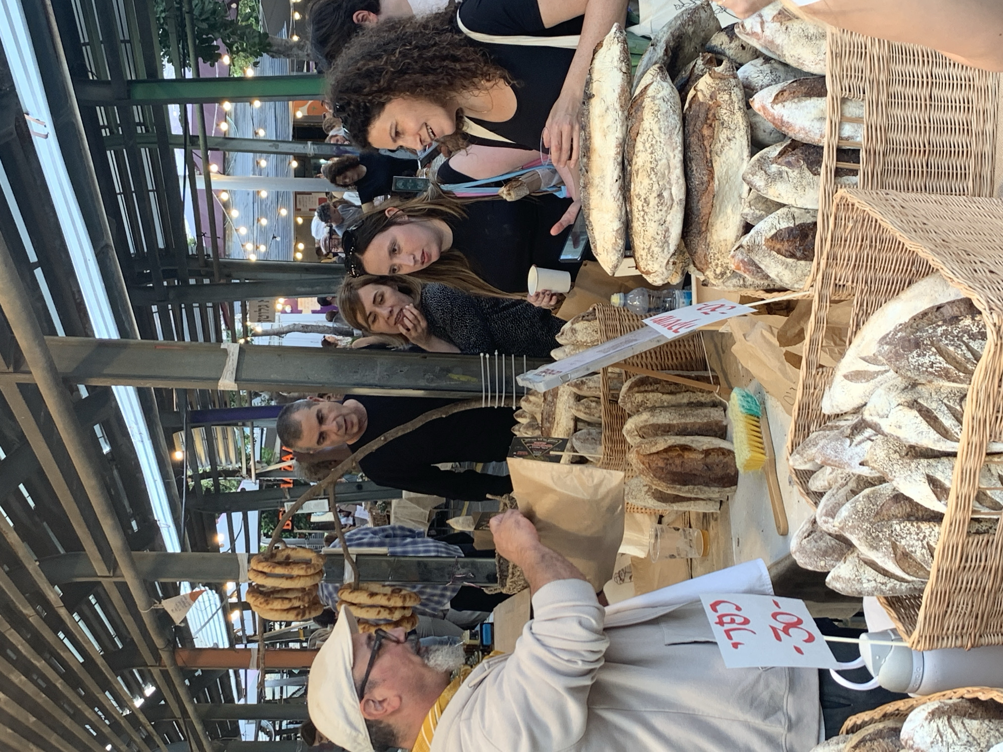 The image captures a lively scene at an outdoor market stall, likely during the evening or night, indicated by the illuminated string lights strung across the overhead metal beam structure. The focus is on a bakery stall, abundant with various types of artisanal bread.
In the foreground, to the left, a male vendor wearing a white cap and a light-colored top is busy handling a brown paper bag, possibly packaging an order for a customer. He has a beard and appears focused on his task. On the opposite side of the stall, several customers are gathered, observing the baked goods. Three women are prominent: one with curly hair smiles warmly as she looks down at the display, another with long dark hair holds a white cup while browsing, and a third, with lighter hair, leans casually on the counter. A man in a black shirt is also visible behind the vendor, looking towards the stall.
The stall itself is filled with a rich assortment of bread, predominantly rustic loaves with floured crusts, displayed in traditional wicker baskets and directly on the counter. There are elongated loaves resembling baguettes, as well as rounder, sourdough-style breads. A unique detail is a string of flatbreads or similar baked items hanging from a wooden branch, enhancing the artisanal aesthetic. A small plastic water bottle and a small blue and yellow brush are also visible on the counter.
A visible white sign with handwritten red text clearly displays "30- שק", indicating a price of "30 Shekels" for an item, consistent with the market's location in Tel-Aviv, Israel. The atmosphere appears pleasant and engaging, with customers actively browsing and interacting at the stall.