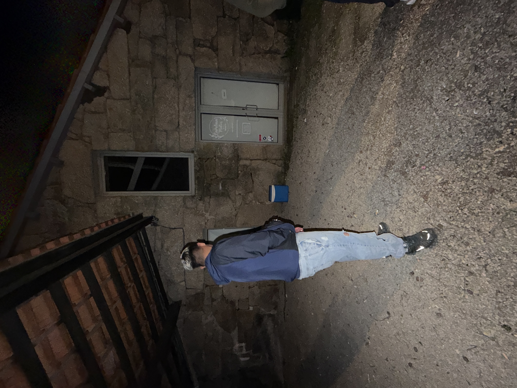 A man with bleached hair, wearing a blue jacket, ripped jeans, and dark sneakers stands on a paved surface in an outdoor area. A stone building with two windows is behind him. The larger window has an advertisement with Portuguese writing. The location is Vila Real, Portugal.