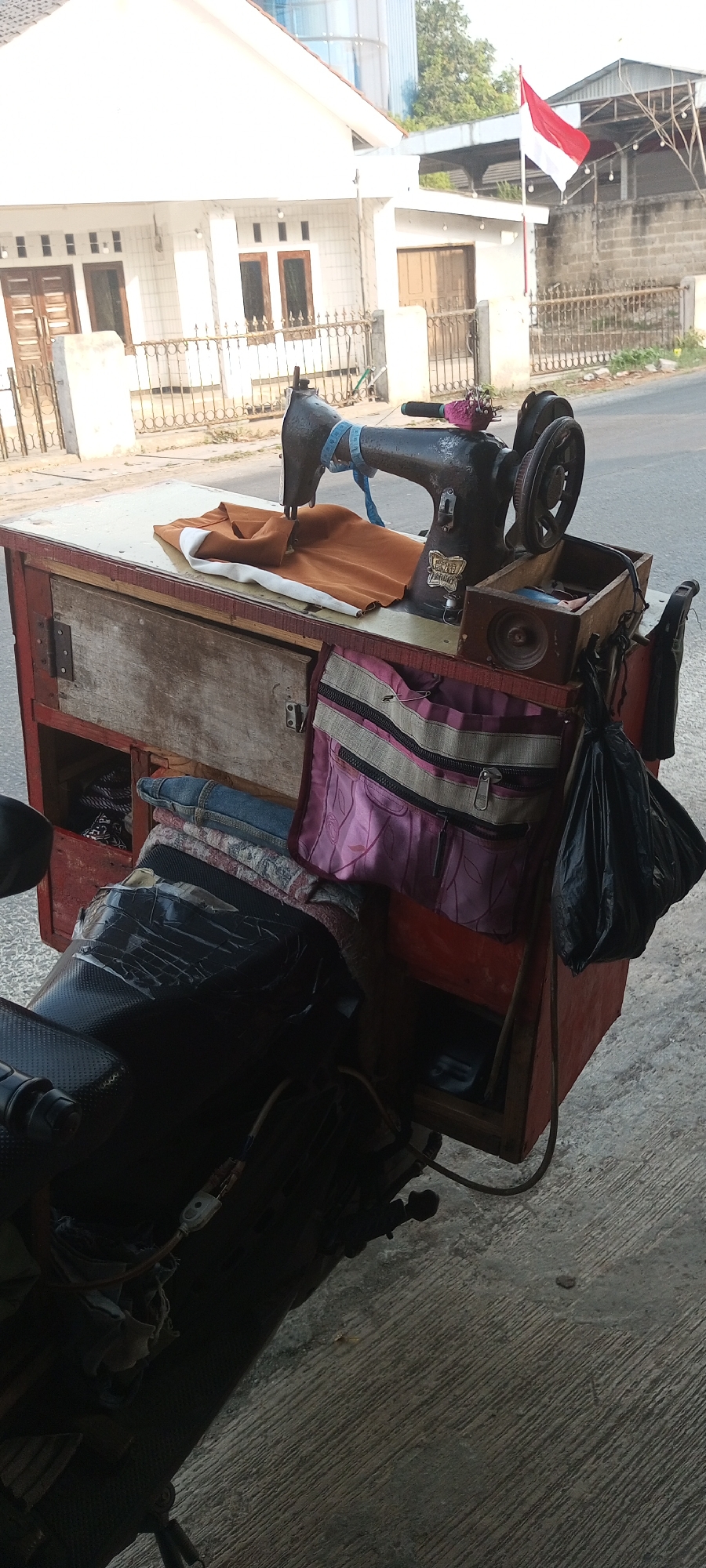 This is a medium shot of a portable sewing machine set up on a red cart, attached to the back of a motorcycle. The cart has a wooden top with a sewing machine on it. To the right of the sewing machine is a small wooden box containing sewing supplies. Hanging from the side of the cart are two fabric bags, one pink with zippers and one black plastic bag. Underneath the pink bag, there are folded clothes, including jeans. The motorcycle seat is covered in black plastic wrap. In the background, there is a white house with a fence in front of it, and a red and white flag flying. The scene appears to be on a street in Setu, Indonesia.