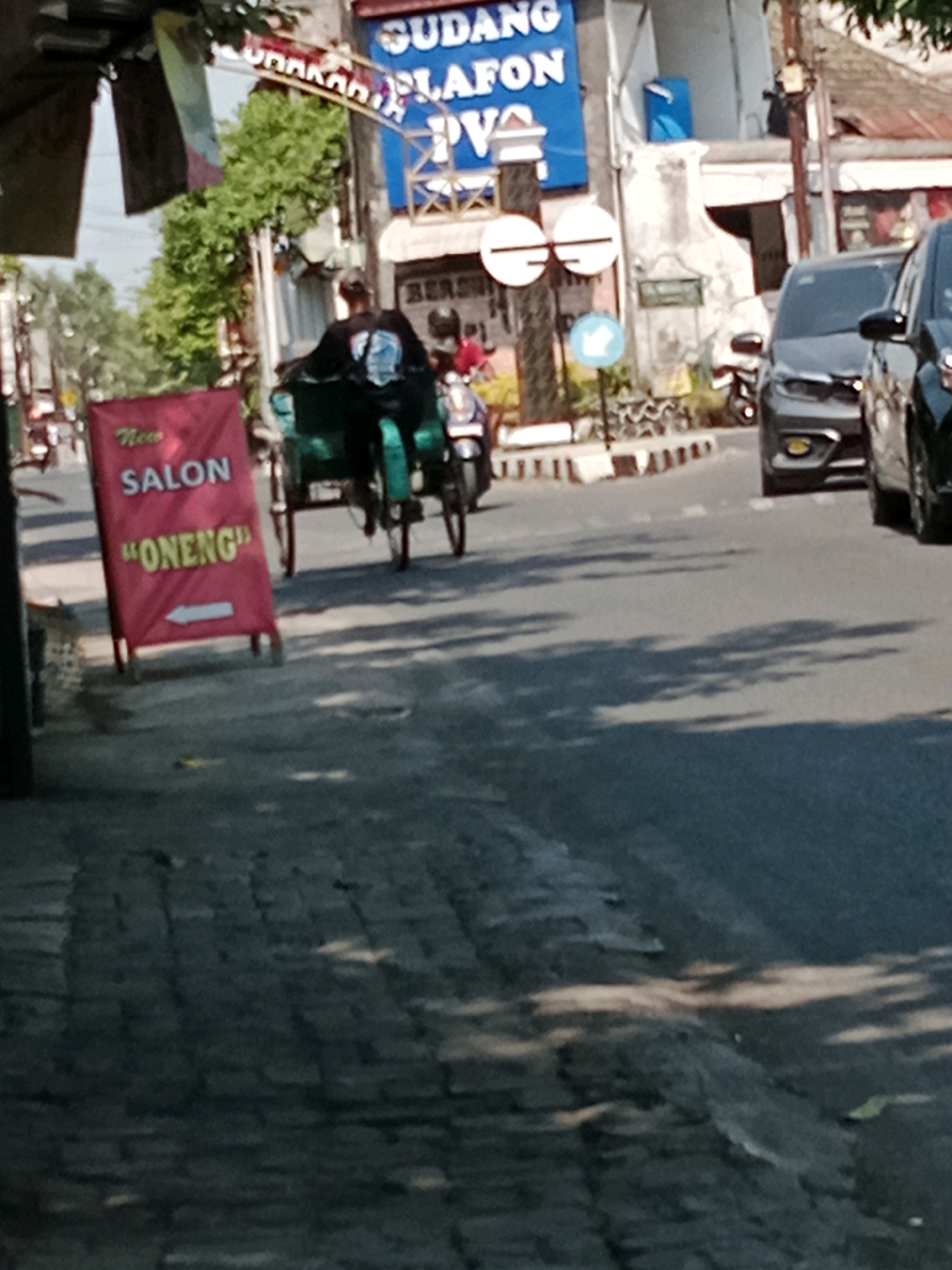 In Banaran, Indonesia, a tri-wheel vehicle, likely a becak, with a rider wearing a dark top and what appears to be a blue and white patterned shirt is stationary on a paved road. The becak's cargo area is a dark green color. To the left of the becak, a red banner with white lettering reads "New SALON" and "ONENG" with a white arrow pointing left. In the background, a building displays a large blue sign with white text that is partially legible as "GUDANG PLAFON" and "PVA". Other vehicles, including a dark-colored car and a motorcycle, are visible on the road. Trees and overhead power lines are also present. The scene is illuminated by natural daylight, casting shadows on the road.