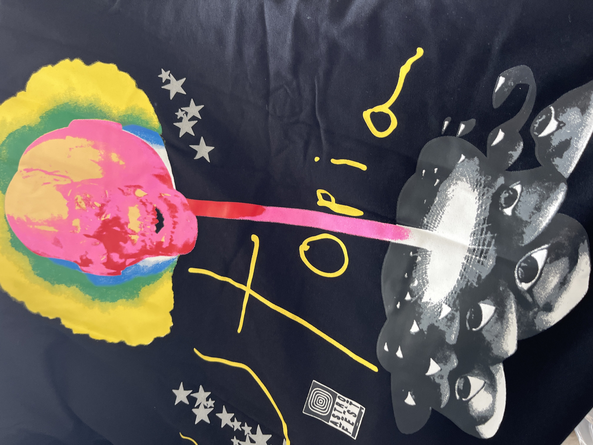 The image shows a dark blue or black fabric or surface with a print design featuring several distinct elements. On the left, there's a brightly colored, abstract skull-like form with yellow, green, blue, pink, and red sections. Emanating from this form is a pink and red stripe. To the right, there's a grayscale image of conjoined or multiple faces with prominent eyes. Additional design elements include yellow scribbled lines forming the word "Rio," scattered white stars, and a small, white rectangular logo with concentric circles and text "Astrid Feses." The specific context or location in Zlatograd, Bulgaria, is not discernible from the image alone.