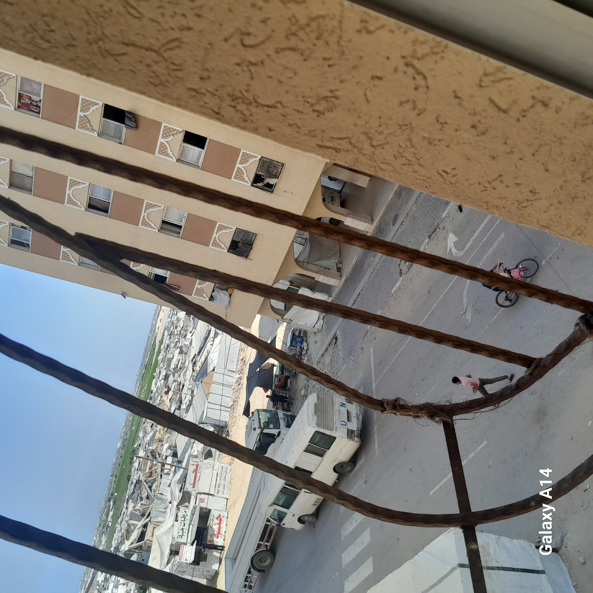 The image captures a scene in the city of Khan Yunis, Palestinian Territories, as viewed from an elevated perspective. In the foreground, weathered, dark brown ropes create a textured barrier, obscuring parts of the view below. To the left, a section of a beige and pink building with multiple windows is visible. Some windows are open, with curtains or clothing hanging out.
Below the building, the urban landscape unfolds. Tents and temporary structures, likely part of a refugee or displacement camp, dominate the middle ground. A white UN marked bus is parked on a paved road, alongside a larger truck. In the distance, a green field contrasts with the densely packed white structures.
On the road, a person is seen cycling, with a pink bag on their bicycle. Further down the road, another person in a pink shirt and grey pants is walking. The road has white lane markings, including an arrow. The overall impression is one of a densely populated area with signs of both temporary housing and ongoing activity.