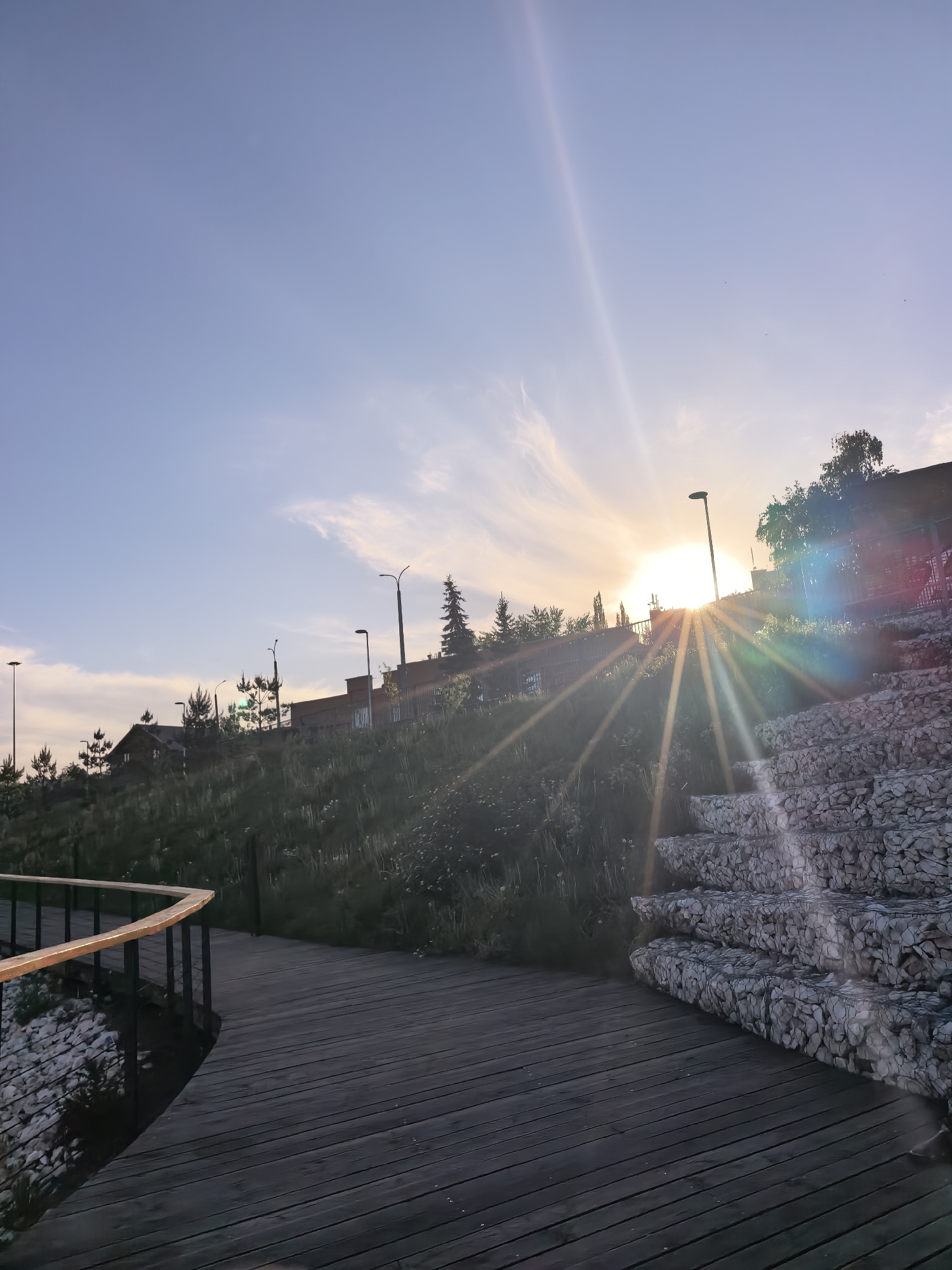 The image shows an outdoor scene in Sterlitamak, Russia, at sunset or sunrise. A wooden boardwalk curves in the foreground, with a railing on one side. To the right, stairs constructed from rock-filled wire cages rise up a grassy embankment. Behind the embankment is a structure with lighting posts. The sun, partially obscured by the structure, casts rays of light across the scene. The sky is blue with wisps of clouds. The vegetation is green and appears unkempt. No people are visible.