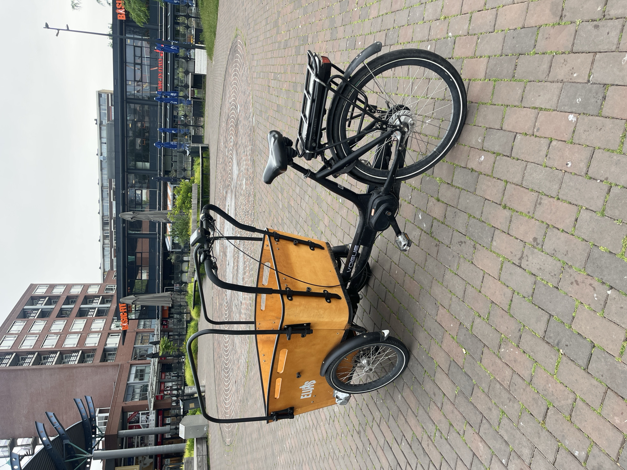 A three-wheeled cargo bicycle with a wooden box is parked on a brick-paved walkway in an urban environment. The bicycle is black with a large, light brown wooden box in the front. The box has the brand name "ELJAS" printed in white on the side. The bicycle has a rear rack and a black seat. In the background, there are multi-story brick buildings with numerous windows. A building with a glass facade and awnings is also visible, along with outdoor seating and umbrellas. The ground is paved with reddish-brown bricks, with some patches of green grass growing between them. The sky is overcast.