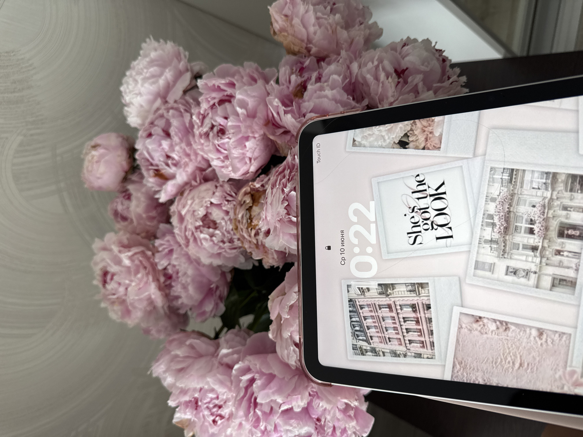 A bouquet of pink peonies is placed behind a tablet device displaying a lock screen with polaroid-style photos of Parisian architecture and a quote. The tablet is held by an unseen person whose hand is partially visible at the bottom left. The background appears to be a textured wall and a surface that reflects light. No specific location in Sochi, Russia, can be determined from the image. The scene is static and indoors.