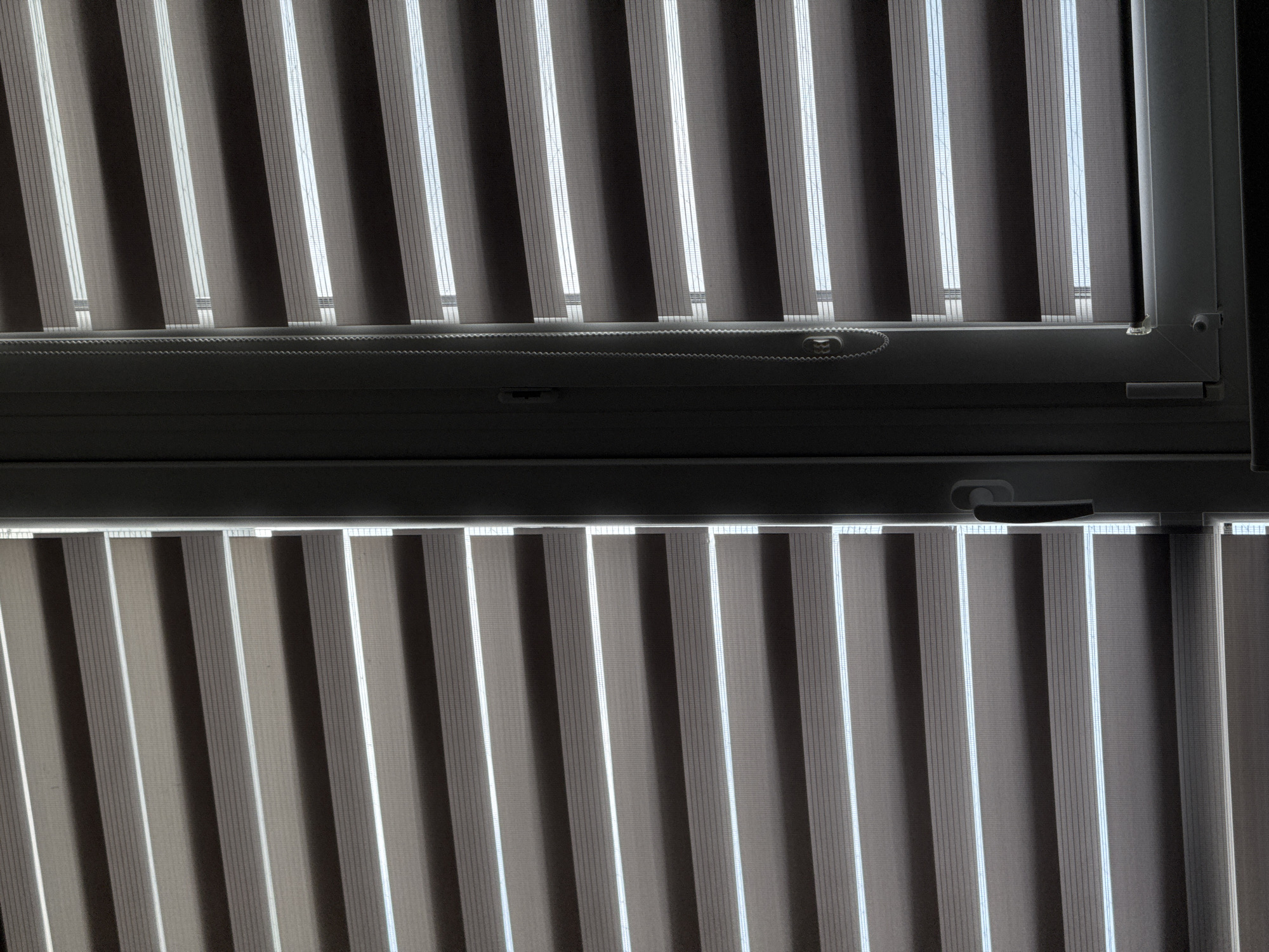 The image displays a close-up view of a window with horizontal blinds. The blinds are a dual-layer type, alternating between translucent and opaque strips, creating a striped pattern. The window frame is visible on the right side, along with a control chain for the blinds and a handle. The lighting suggests that it is daytime, with light filtering through the translucent sections of the blinds. The scene is indoors, looking out through a window. There is no indication of the specific location within Bratislava, Slovakia.