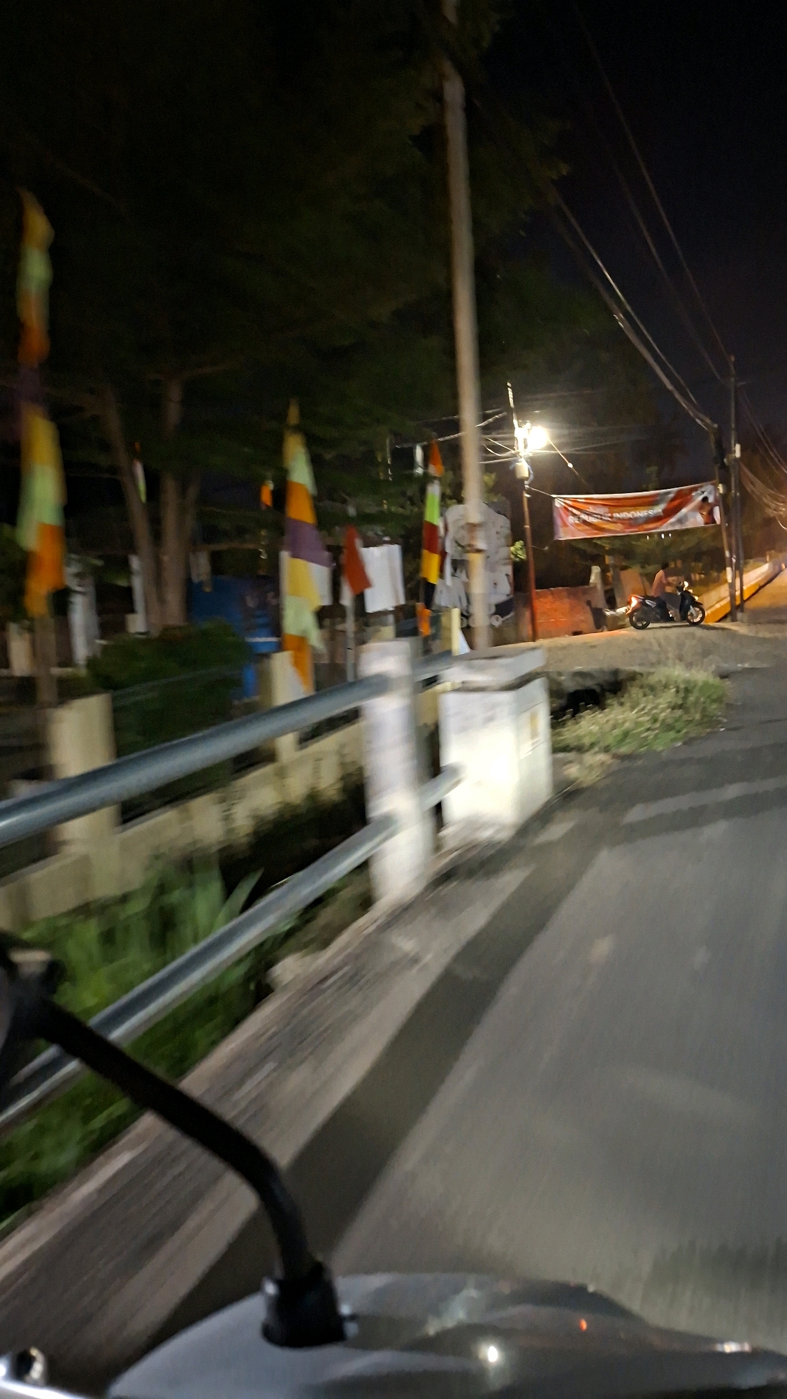 The image, captured at night in Lhokseumawe, Indonesia, depicts a scene from the perspective of a moving vehicle, likely a motorcycle, as indicated by the presence of a mirror in the foreground. The road is paved and appears wet, reflecting the ambient light. On the left side of the road, a concrete barrier with metal railings runs parallel to the path, bordering a channel with green vegetation. Beyond the barrier, several colorful flags, predominantly orange, yellow, green, and purple, are displayed on poles, suggesting a festive or event-related setting. Trees with dark foliage are visible behind the flags. To the right of the road, a red and white banner with text, possibly "REPUBLIK INDONESIA," is hung across a street pole. A person is riding a motorcycle under the banner, illuminated by a streetlamp. The overall environment suggests a public space or street within the city, possibly near a residential area or a location hosting an event.