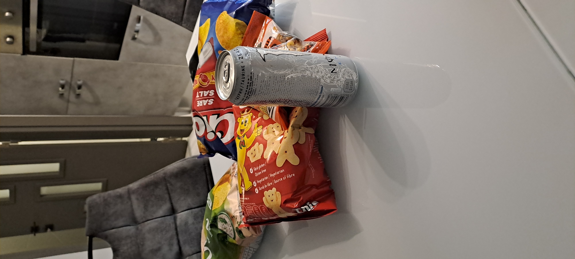 This image captures an indoor scene, likely in Burgas, Bulgaria, showing a collection of snacks and a drink placed on a white surface, possibly a mini-fridge or a table. The primary subjects are several bags of chips and a silver energy drink can, arranged somewhat haphazardly.
In the foreground, a silver Monster energy drink can is prominent, lying on its side. Visible text on the can includes "ТАУРИН/TAURINE + ZERO" and "ZERO SUGAR," indicating a sugar-free taurine-based beverage. Below the can, a red snack bag features cartoon gingerbread-man-like characters. Text on this bag specifies "Fără gluten / Gluten free," "Vegetarian / Vegetarian," and "Sursă de fibre / Source of fibre," suggesting a health-conscious snack. Part of a brand name, "Unis," is also visible. To the left of the energy drink, a blue and red bag of chips or crisps is seen, with the words "SALT" and "SARE" (Romanian/Italian for salt) clearly visible, along with the brand "Qio." Another green bag of snacks is partially visible at the bottom left.
The background shows interior elements. On the left, there's a grey, textured cabinet or wall with small metallic handles and what appears to be a dark reflective surface above it. Below this, a dark grey upholstered chair or sofa is partially visible. The lighting suggests an indoor setting, and there's no indication of the time of day or outdoor weather. The multi-lingual text on the snack packaging (English, Romanian/Bulgarian, Cyrillic) aligns with the specified location context of Burgas, Bulgaria.