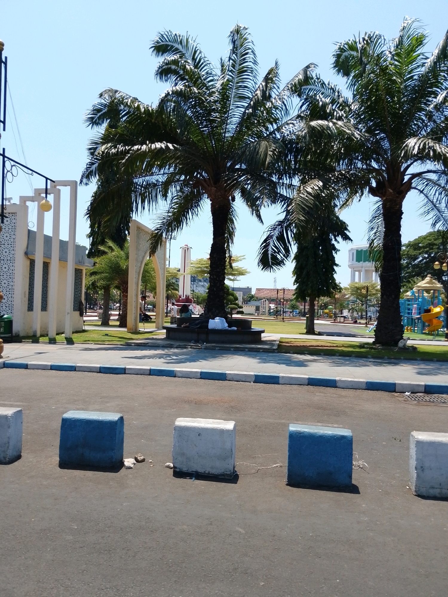 The image depicts a sunny day scene in Pasuruan, Indonesia, likely at a public park or plaza. In the foreground, there are three concrete barriers on an asphalt road, two of which are painted blue and white. Beyond the road, a green lawn is visible, bordered by a pathway with blue and white curbs. In the background, several tall palm trees dominate the landscape, with their fronds reaching towards a clear blue sky.
To the left of the palm trees, there is a modern white building with decorative geometric patterns on its facade and tall vertical pillars. An ornate archway, also white and decorated with geometric patterns, stands slightly to the right of the building, framing a view of the park.
Further into the park, a children's playground with colorful slides and climbing structures is visible. A tall white water tower with a green top stands in the mid-ground, and a red and white striped monument is also present. Several people are scattered throughout the scene, some sitting on benches, others appearing to walk. The overall impression is that of a well-maintained public space in a tropical or subtropical urban environment.