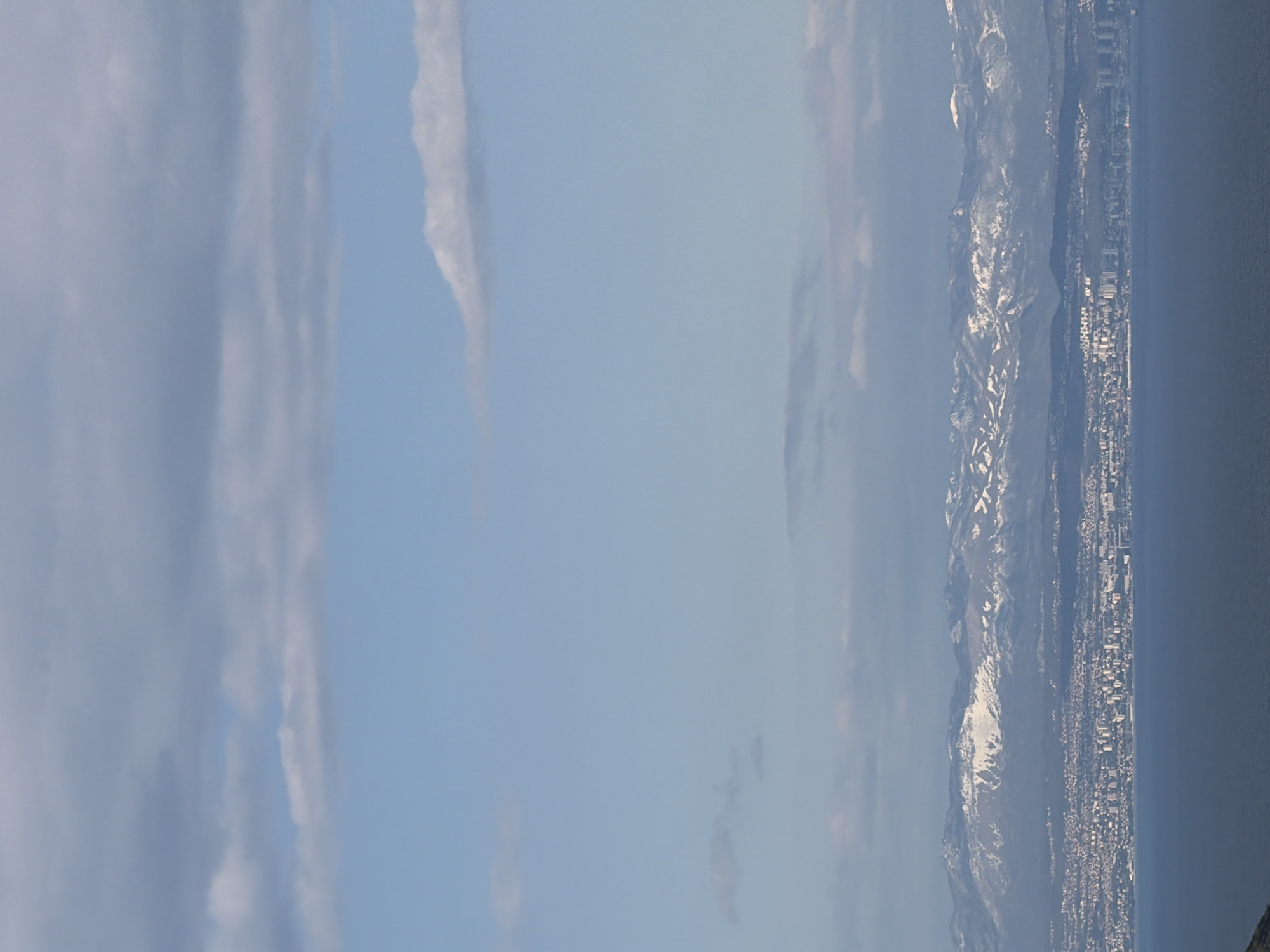 The image shows a view of Zagorje, Croatia from an elevated perspective. The sky is predominantly blue with scattered clouds. In the distance, a mountain range is visible, partially covered with snow. The city stretches along the coastline, with buildings and structures densely packed together. The sea is dark and calm. The overall scene depicts a landscape blending urban and natural elements.
