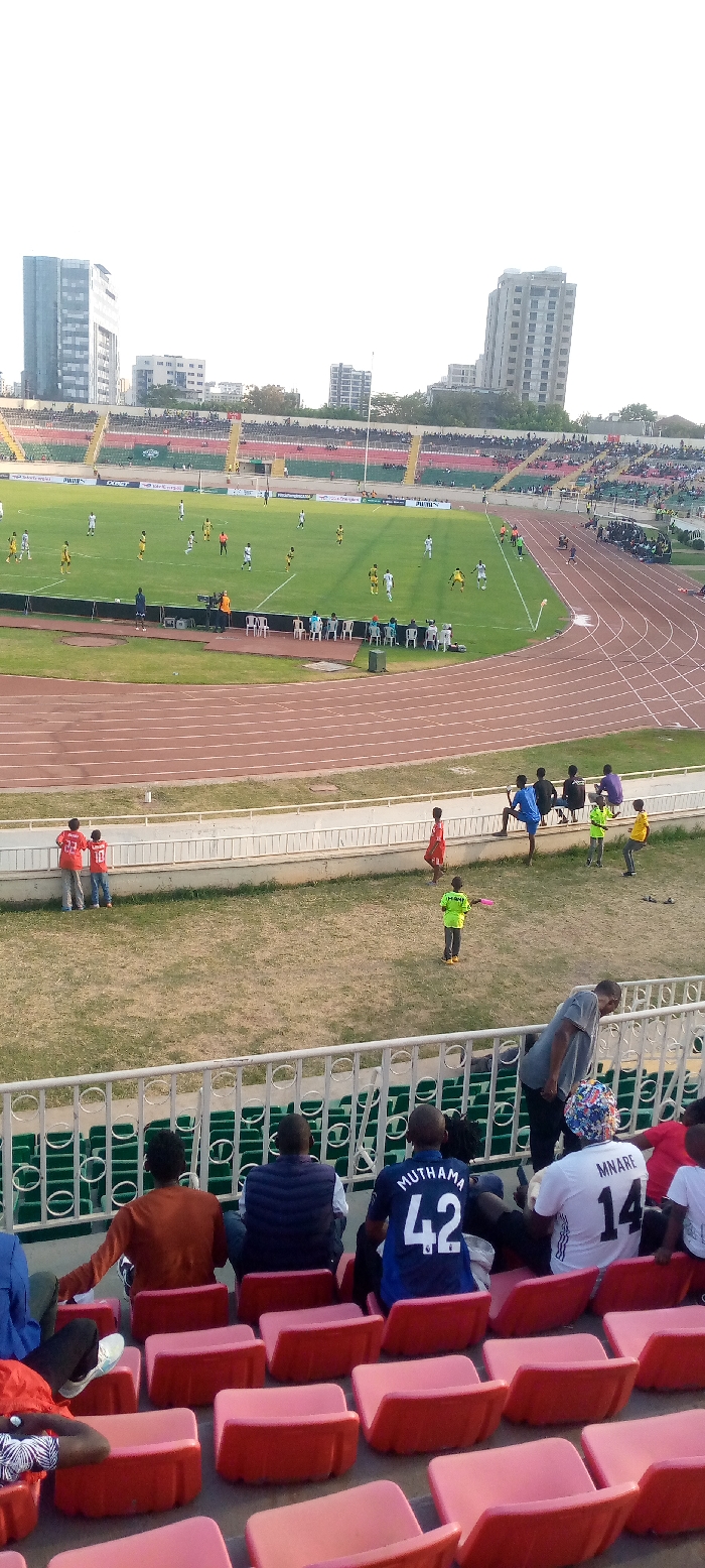 An elevated view shows a stadium during a soccer game. A track encircles the field. Spectators occupy the stadium seating. Buildings are visible in the background. In the foreground, stadium seats are mostly unoccupied, some with seated spectators. People are standing in the open area between the track and the seating. A mix of children and adults are scattered throughout this space.