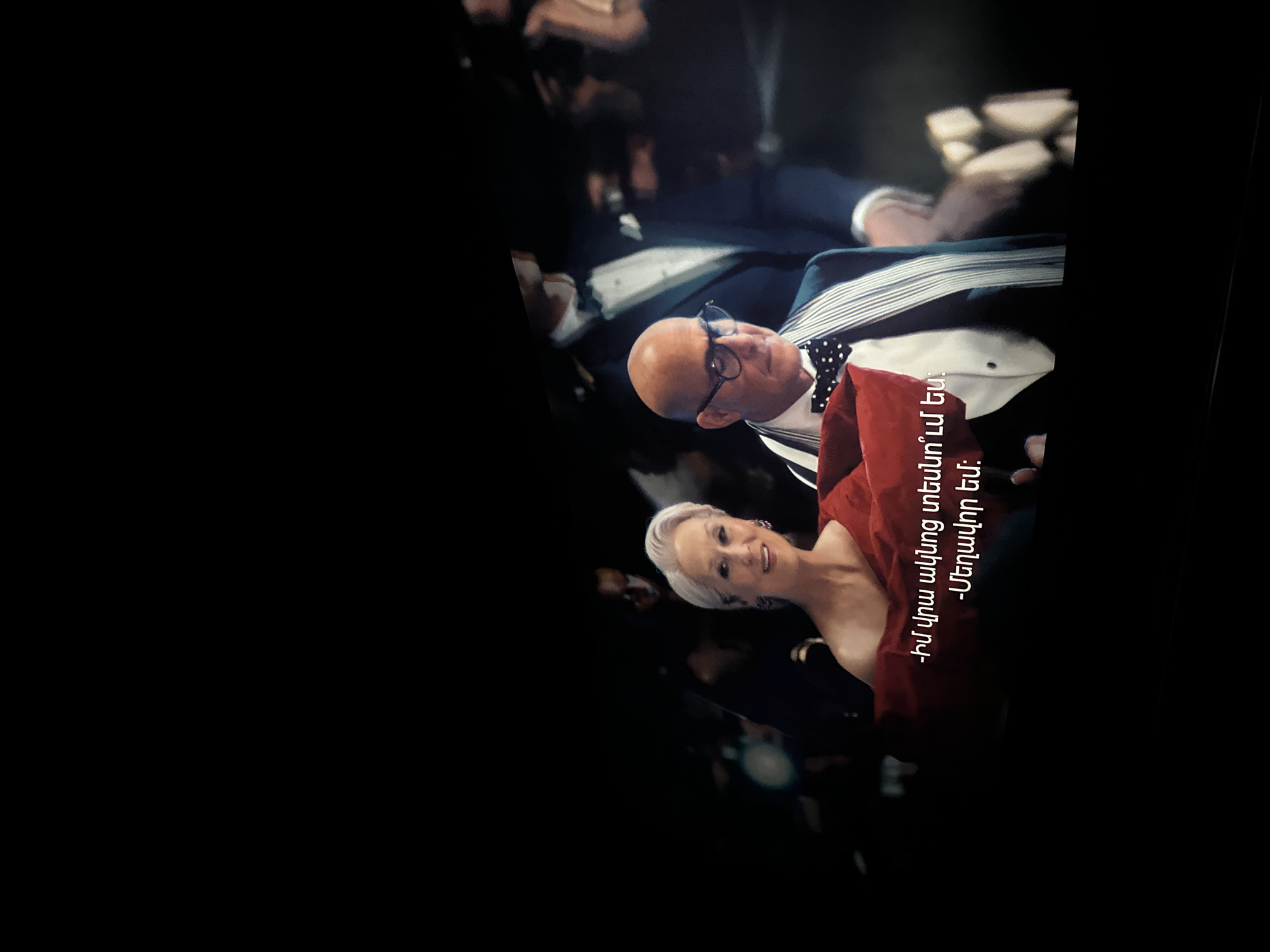A photo captured indoors displays a man and a woman posing for the camera at an unknown event. The woman, wearing a red dress, smiles, while the man, clad in a black suit with a white shirt and a black bow tie, gazes into the distance. Overlaying their image is white Armenian text. The setting appears to be a gathering or ceremony, but the image gives no specific indication of Yerevan, Armenia.