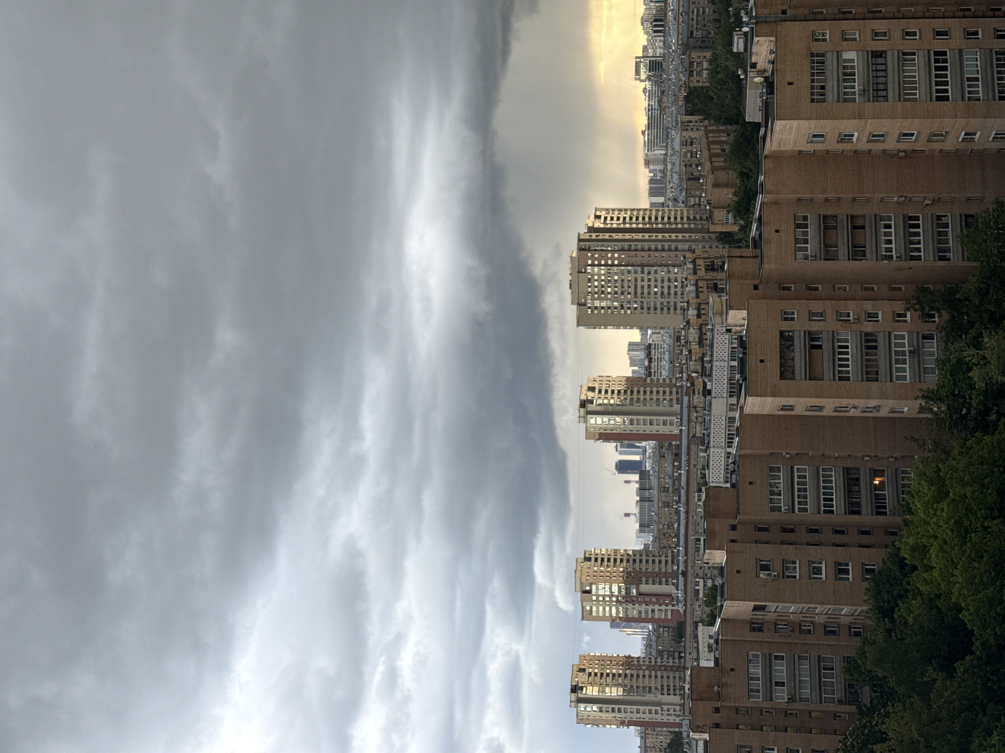The image captures a cityscape in Moscow, Russia, marked by a dramatic weather divide. Dominating the left side of the frame are imposing, dark storm clouds, suggesting an imminent downpour. In contrast, the right side of the image is bathed in a warmer, brighter light, illuminating rows of residential and commercial buildings.
The architecture is typical of Soviet-era construction, featuring large, blocky apartment buildings with rows of windows. Some buildings show signs of more recent renovation, while others appear older and more utilitarian. Interspersed among the buildings are patches of green trees, indicating parks or residential landscaping.
There are no people immediately visible in the image. The scene conveys a sense of transition, as the city prepares for an approaching storm while still enjoying a moment of sunlight.