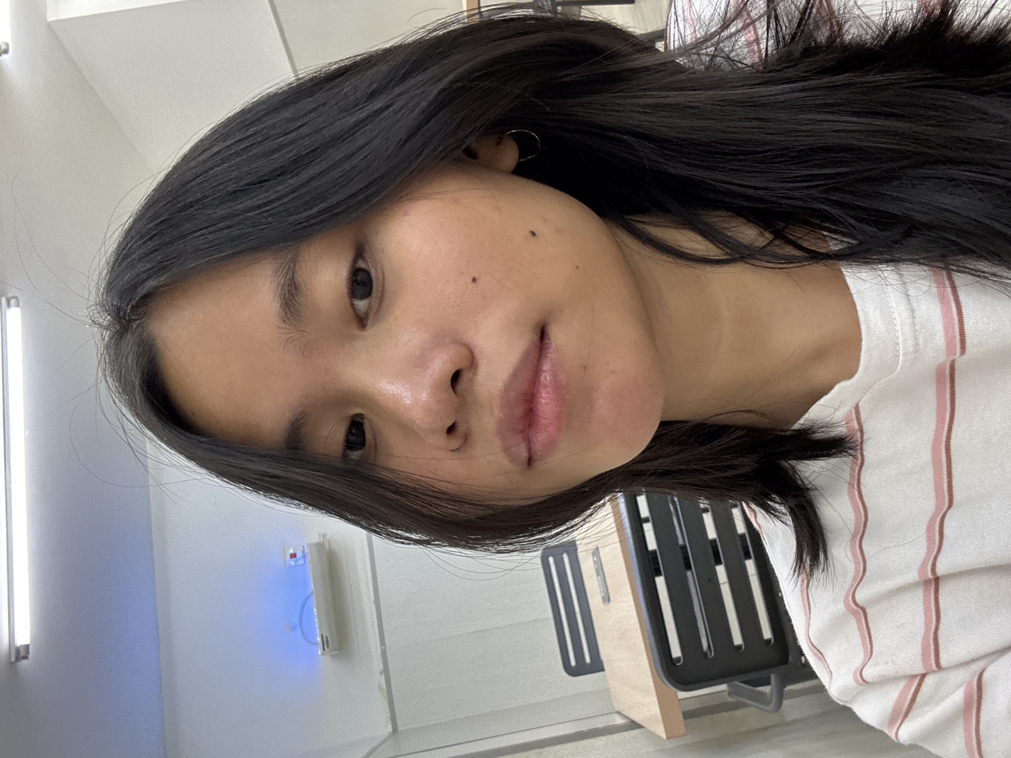 The image is a close-up, eye-level, selfie-style photograph of a young East Asian woman. She has long, dark hair that falls around her face and shoulders. Her skin is light-toned with a few freckles and two distinct moles visible on her right cheek. She is wearing a white t-shirt with thin pink and brown stripes. A small gold hoop earring is visible in her right ear.
In the background, there is a blurred view of an indoor environment that appears to be an office or study area. A light-colored table and a dark chair with a slatted back are visible to the left of the subject. The wall behind her is white, with a light fixture and what appears to be a power outlet with blue light above the table. The overall lighting suggests an indoor setting. The location is specified as East Jakarta, Indonesia.