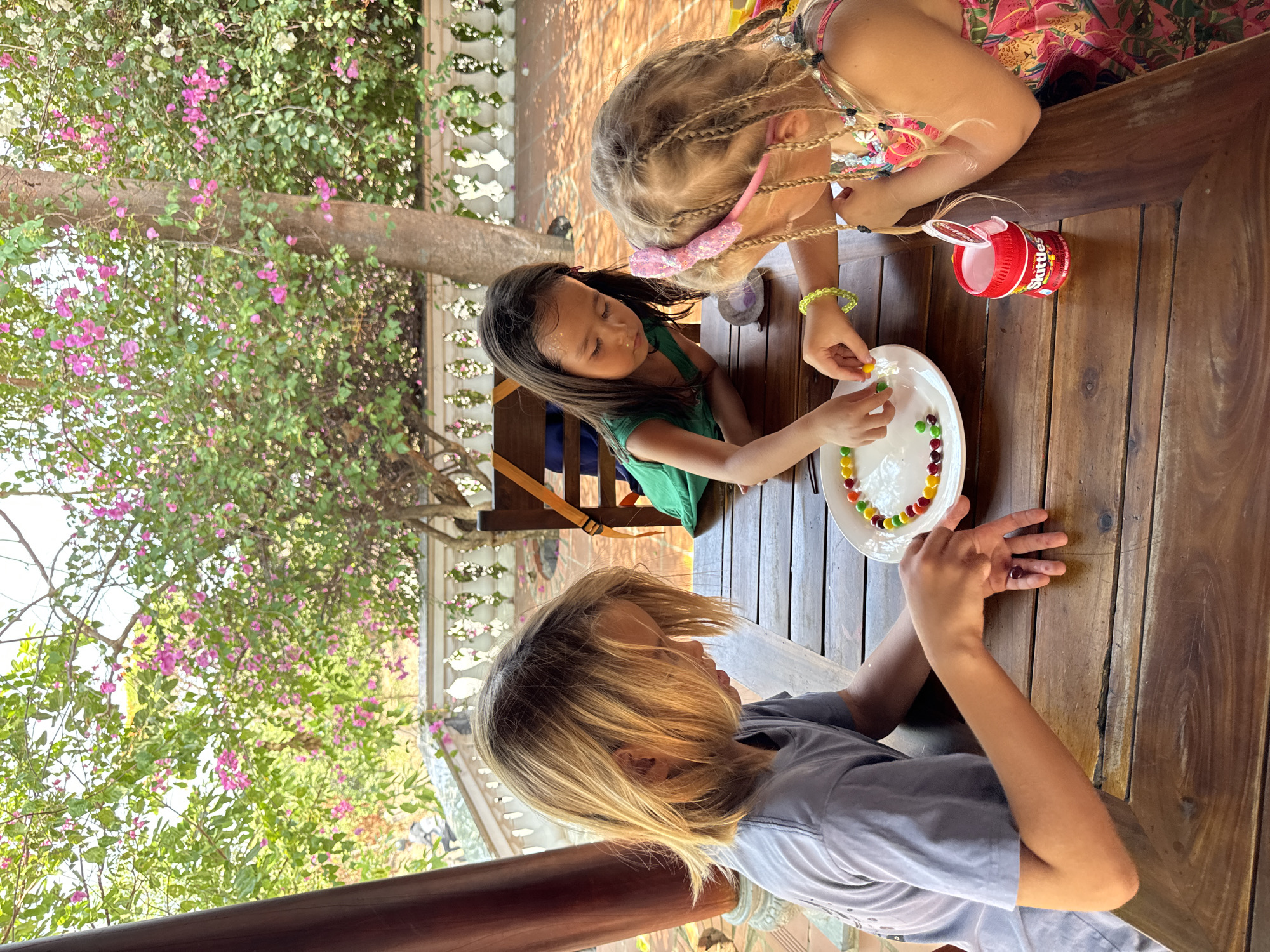 Three children are seated around a wooden table, engaged in an activity involving Skittles candy. A girl with dark hair wearing a green top sits next to a fair-haired girl with braids and a pink headband, both focused on arranging the candies on a white plate. A third child, with short blonde hair and a grey shirt, sits across from them, participating in the activity. A red Skittles container is on the table. Outside, a tree with pink flowers and green foliage is visible, suggesting the scene is set outdoors, possibly on a patio or deck. The setting is in Mũi Né, Vietnam.