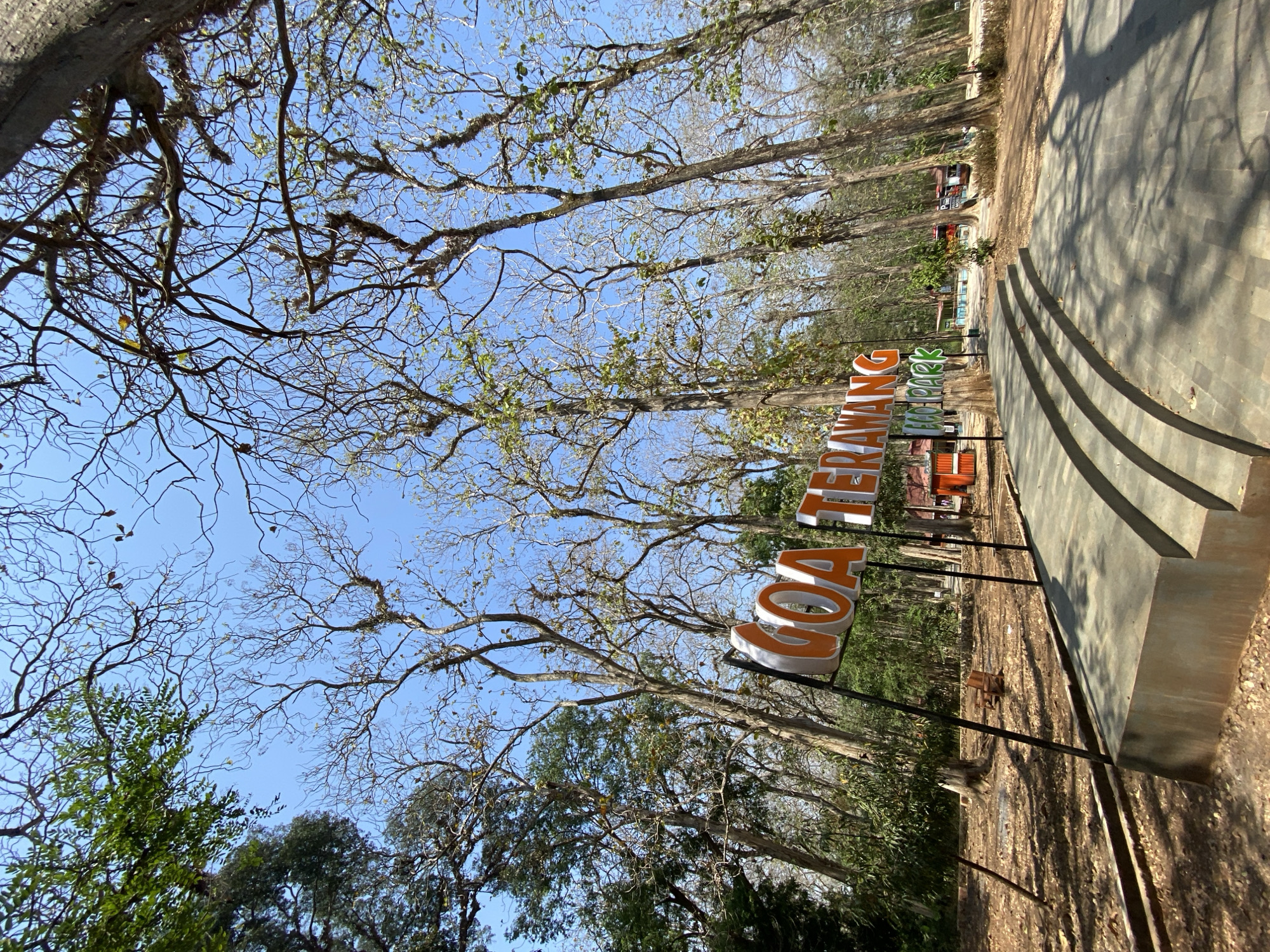 This is a low-angle, full outdoor shot of a park entrance. The foreground features a concrete ramp with three curved steps leading up to a large, three-dimensional sign. The sign is orange and white and reads "GOA TERAWANG ECO PARK". The letters are tall and blocky, with orange faces and white edges. The sign is supported by black metal poles.
Behind the sign, the ground is covered in dry leaves and dirt. There are several tall, bare trees with thin branches reaching up towards a clear blue sky. Some of the trees have a few green leaves. Further back, there are more trees, some of which are densely leaved and green, forming a backdrop. Sunlight casts dappled shadows on the ground and the stairs.
In the middle ground, there are a few small structures. One appears to be an orange building, possibly a kiosk or information booth. Another structure, farther back, is red and appears to be a vehicle or cart. There are a few people or figures visible in the distance, but they are too small and indistinct to identify. The overall impression is of a natural, somewhat arid park environment on a sunny day.