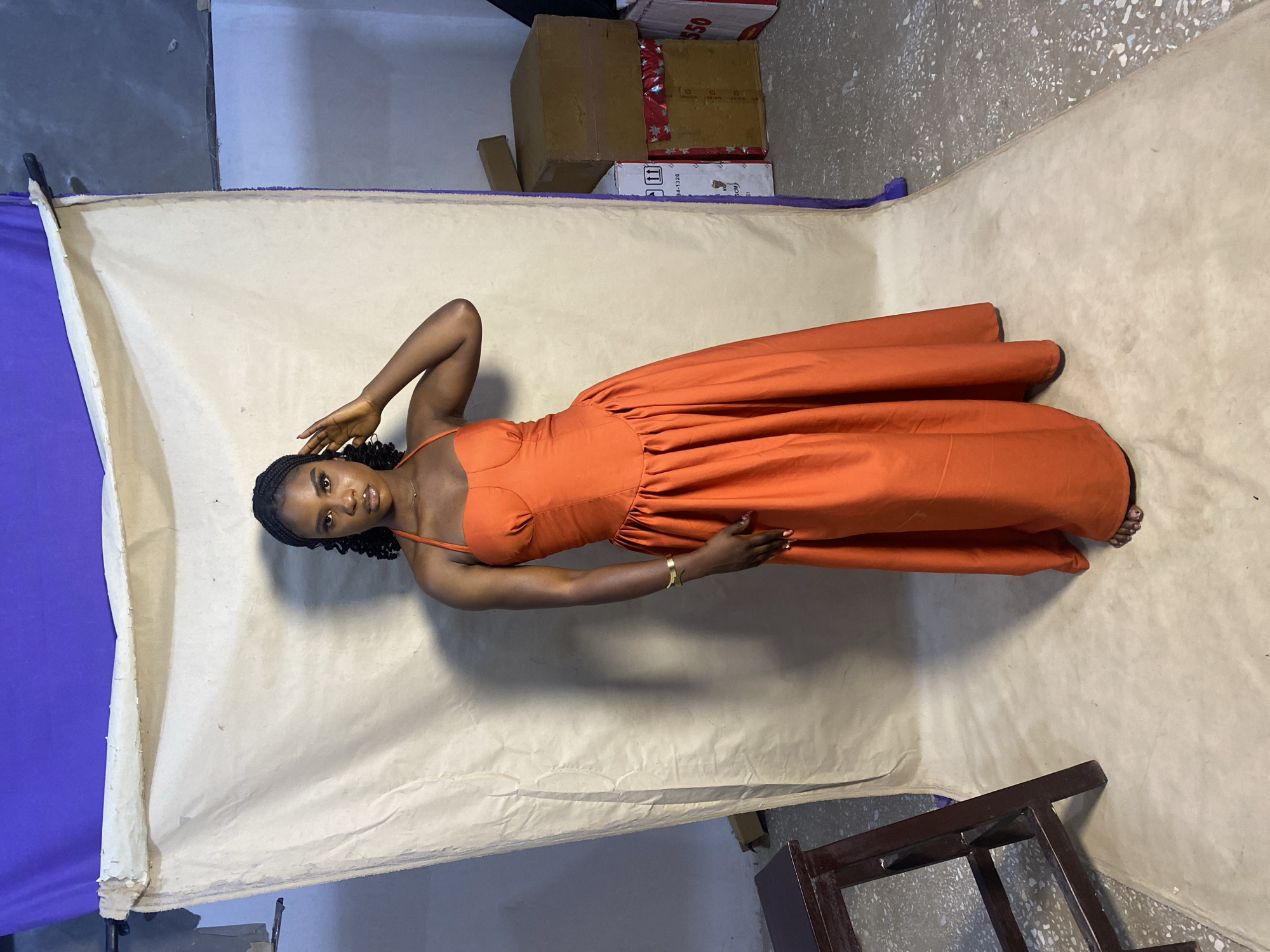 A dark-skinned woman is lying on her side in a long, bright orange dress on a tan backdrop with a purple-covered stand to the left. Her head is oriented toward the left with her right hand on her head and her left hand resting on her thigh. Her braids are visible. Boxes are visible near the top of the frame. A partial chair is visible on the bottom right. The image was likely taken in a studio setting in Jos, Nigeria.