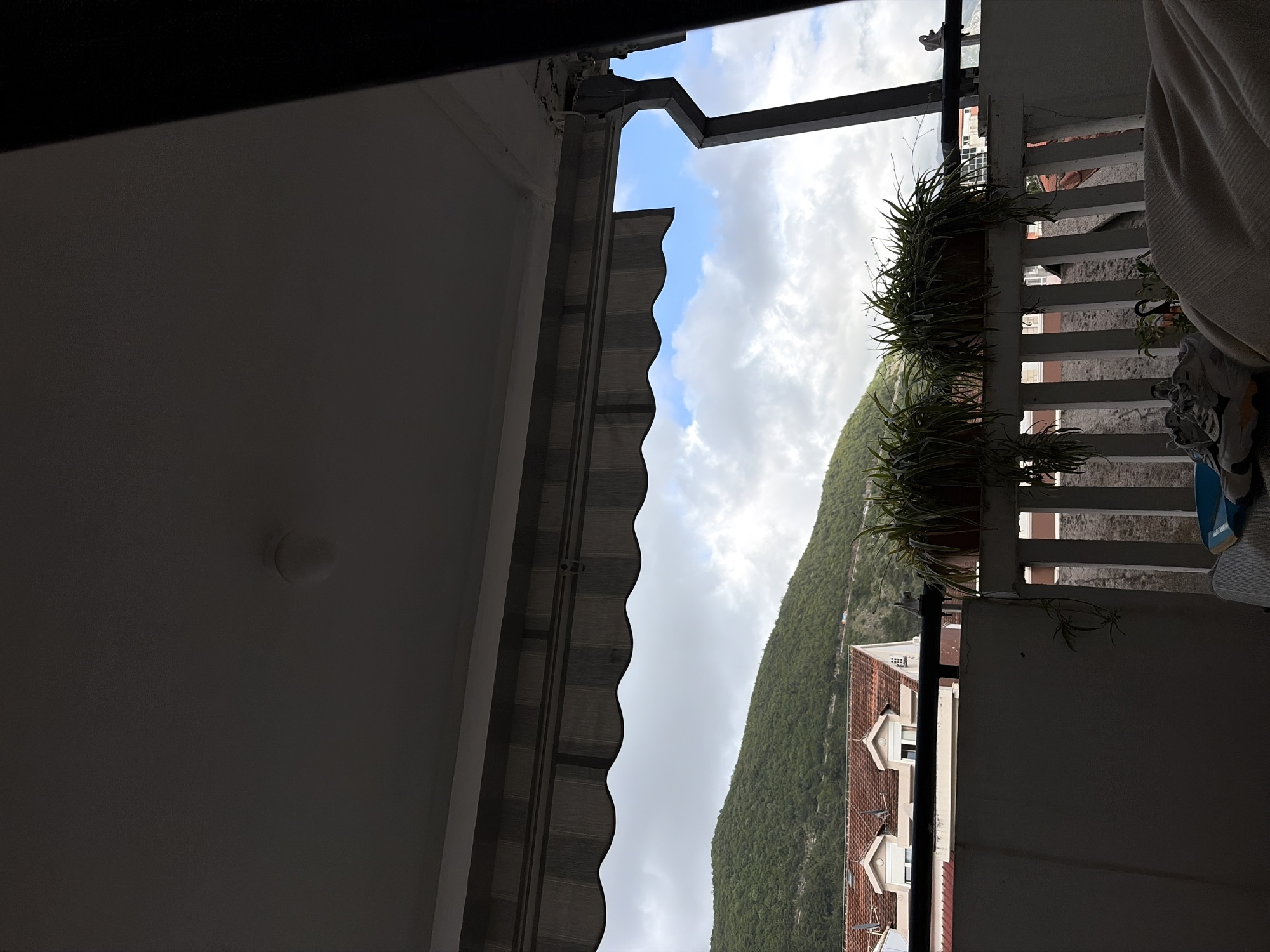 The image, taken from inside a building, captures a partial view of a city landscape, likely in Budva, Montenegro, given the mountainous terrain and red-tiled roofs characteristic of the area. The perspective is from a balcony or window, with a white-painted wall and ceiling visible on the left side of the frame. A gray awning extends from the building's edge. Through the opening, one can see a mountain covered in green vegetation, partially obscured by cloudy skies. A portion of a building with a red-tiled roof is visible at the foot of the mountain. On the right side, a white balcony railing with plants hanging in pots is partially visible, along with draped white cloth and what appears to be clothing. The composition is slightly rotated, and the lighting is somewhat subdued.