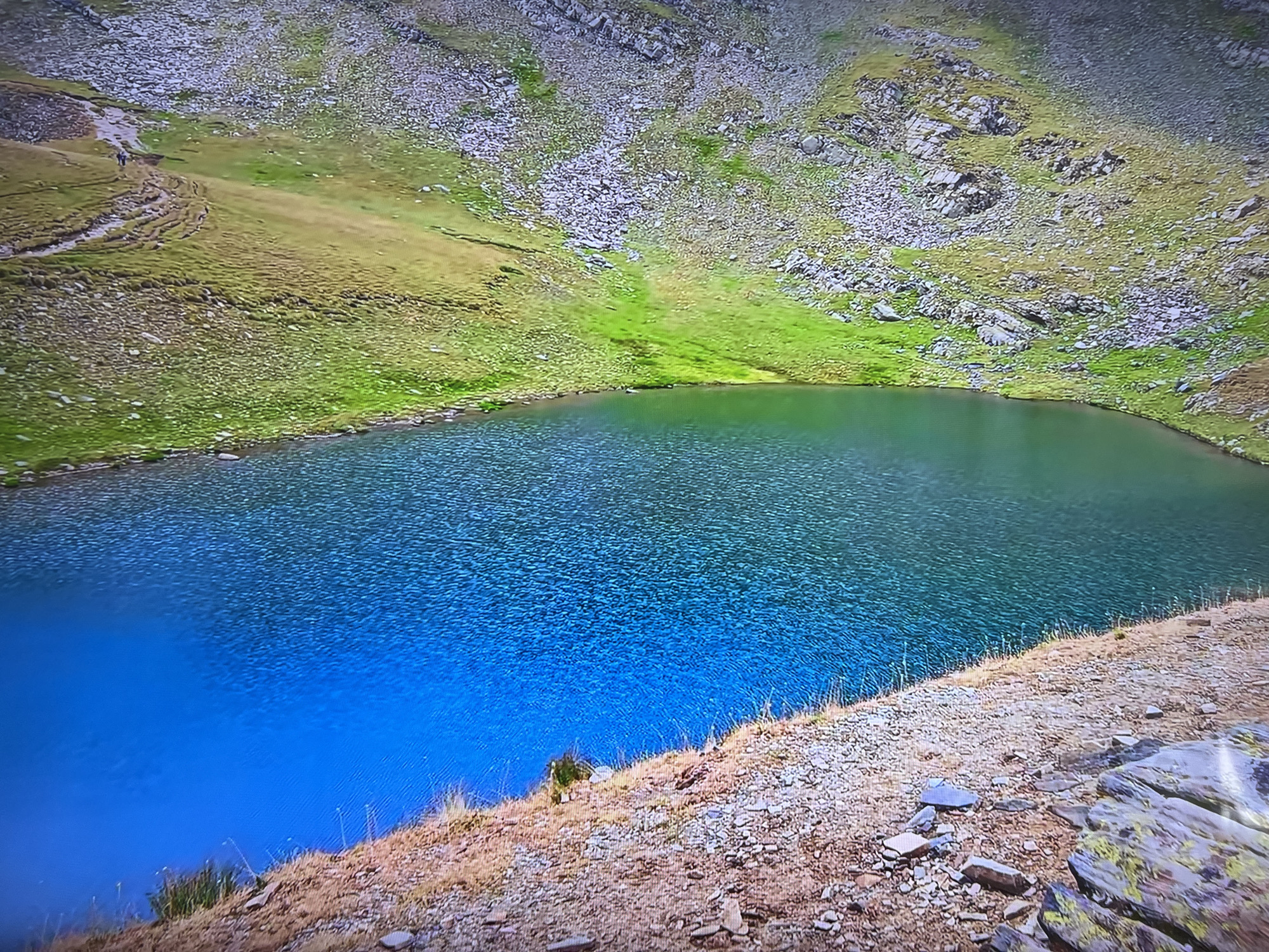 The image depicts a lake surrounded by grassy and rocky terrain. The water color transitions from a deep blue in the foreground to a darker green towards the center and back of the lake. The surrounding land is a mix of green grass and rocky areas, transitioning to larger rocks and scree higher up the hillside. A hiking path is visible on the left side of the image. The location is Unknown, Unknown. No people or interactions are distinctly visible besides the path suggesting human presence.