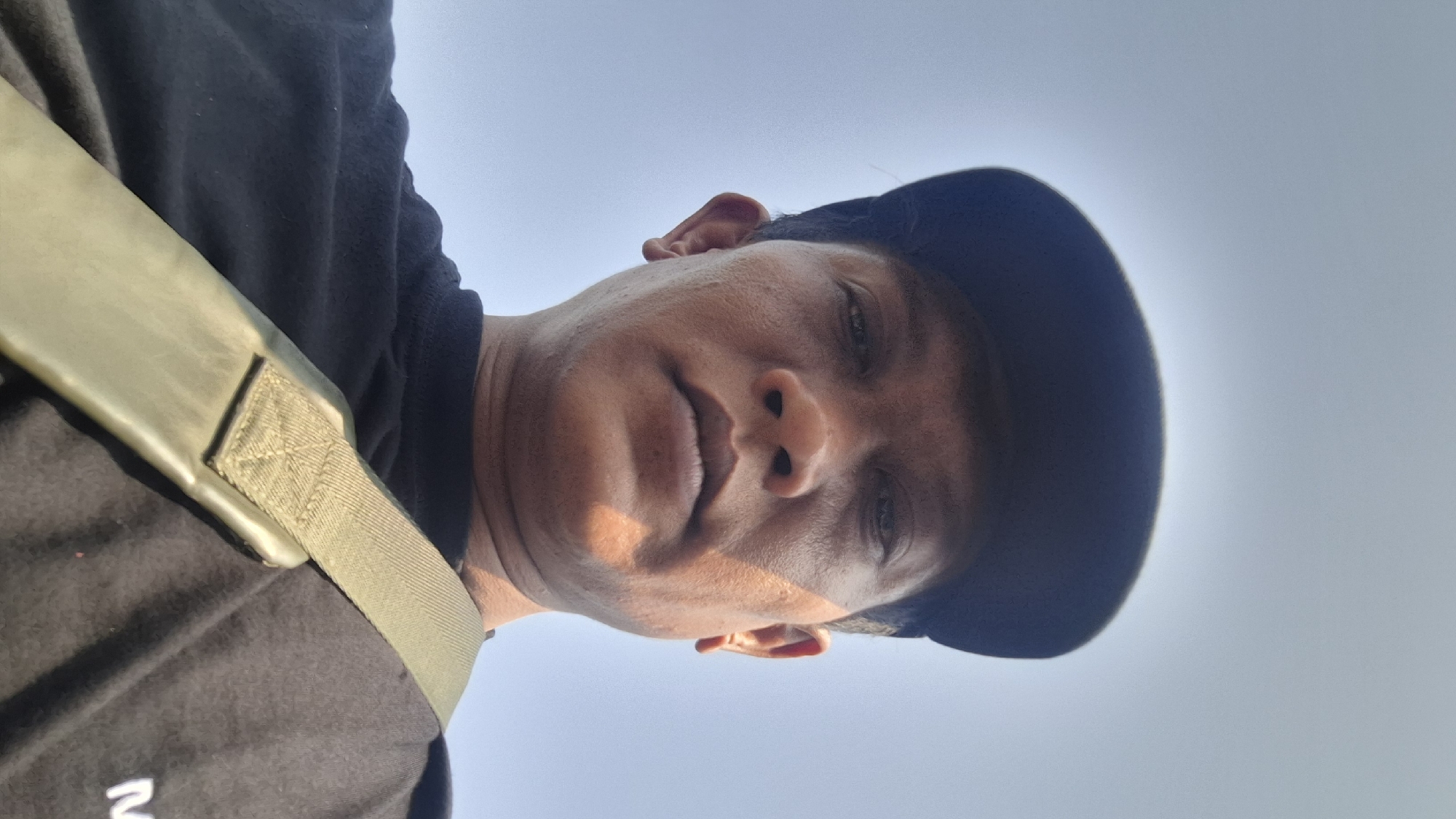 A low-angle selfie taken outdoors shows a person wearing a black t-shirt and a dark baseball cap. The person's face is angled down towards the camera, with their head tilted back. The lighting suggests it is daytime, with sunlight casting shadows on the right side of their face. The background is a clear, pale blue sky. A green strap crosses the person's chest. The image appears to be taken in Rahong, Indonesia, based on contextual information.
