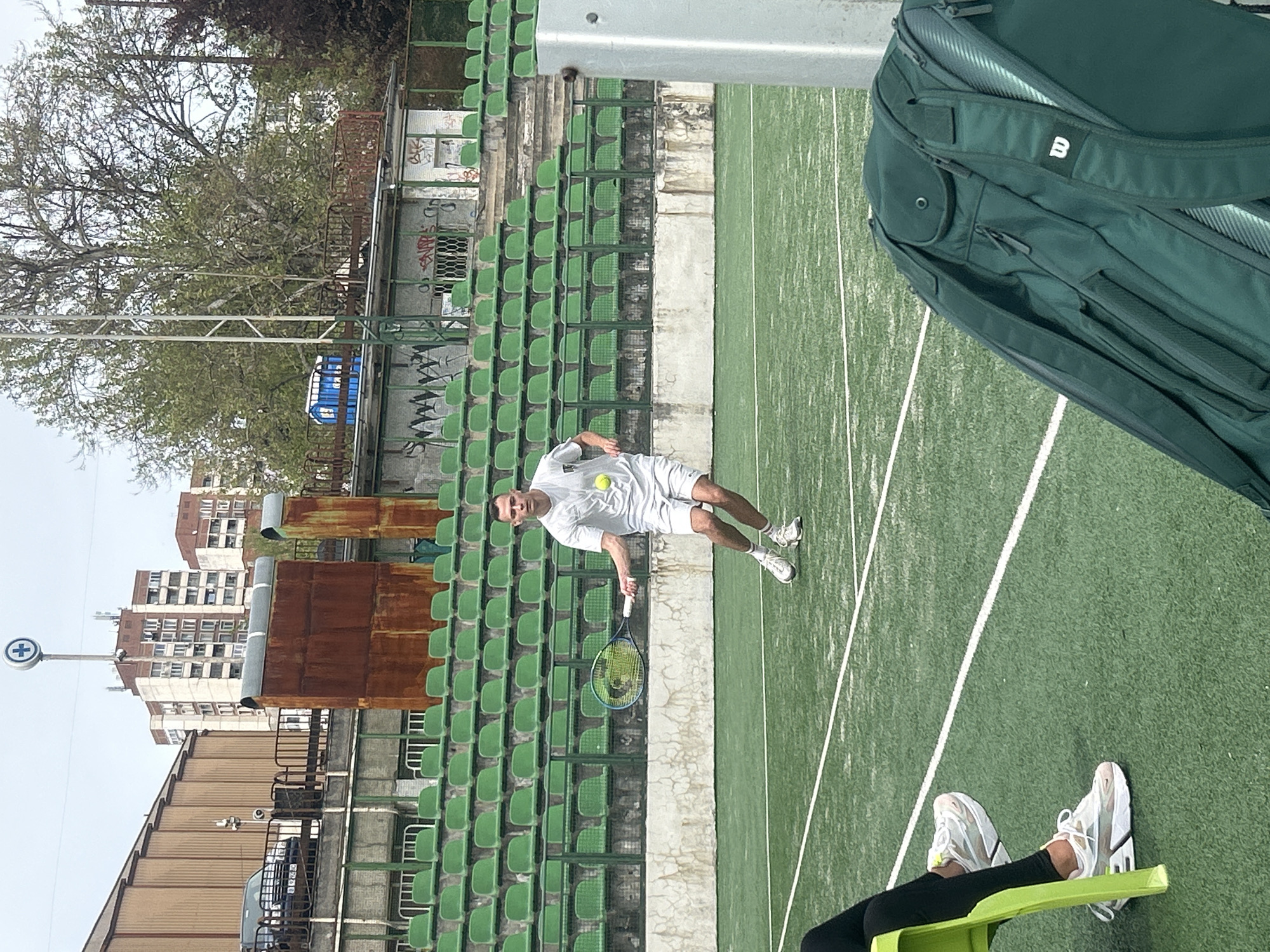 The image shows a man in a white athletic outfit playing tennis on a green court with white lines. He is in the middle of serving the ball with his tennis racket raised. Behind him are rows of green stadium seating that appear old and weathered. A tall, rusted structure is also visible behind the seating. In the background, there are residential buildings. In the foreground, a person's legs and shoes are visible, suggesting someone is watching the match. A green tennis bag is also present, partially obstructing the view on the right. The location is Veliko Tarnovo, Bulgaria, as indicated by the distinctive "+" sign on a pole near the buildings.