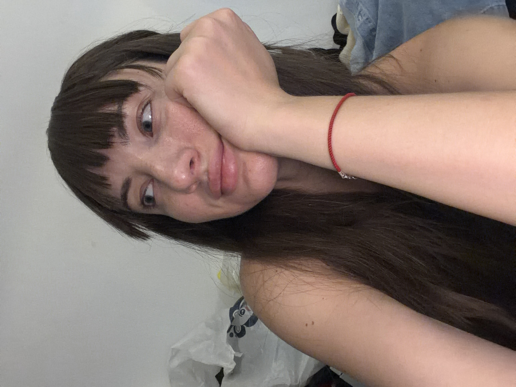 A woman with dark hair and bangs leans her head on her hand, looking away from the camera. She is wearing a red string bracelet. The background is a plain wall with some white plastic bags and a denim garment. There is no indication of this image being taken in Sharm El Sheikh, Egypt.