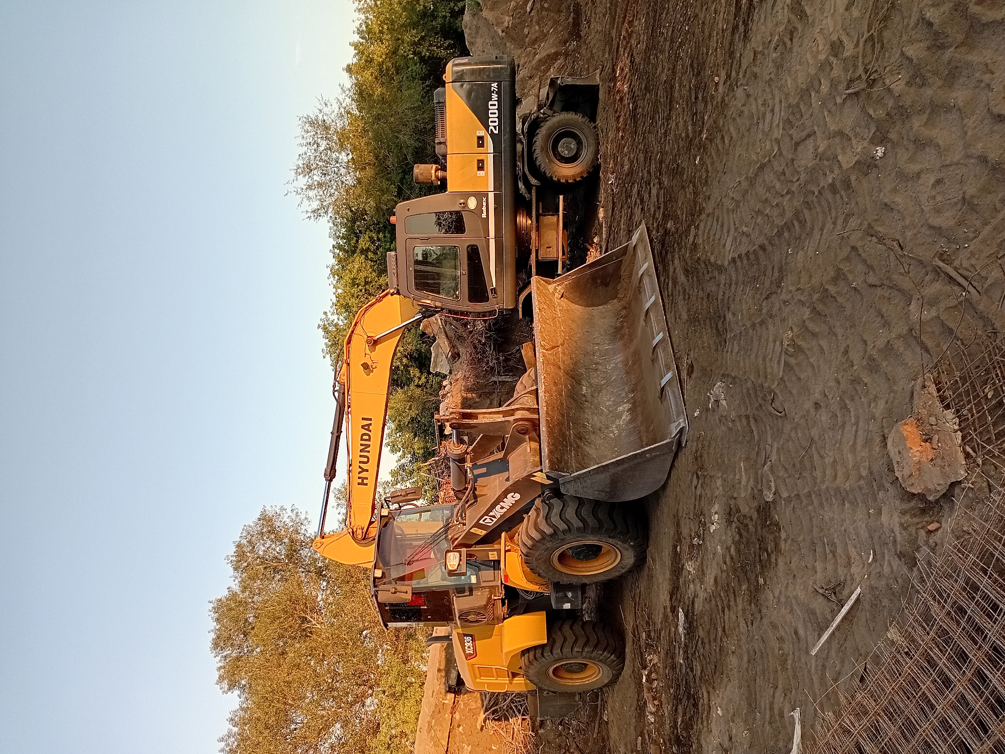 Two construction vehicles, a yellow Hyundai excavator and a yellow XCMG wheel loader, are parked on a dirt construction site under a clear, pale blue sky. The excavator, on the right, has the model number "2000W-7A" visible on its side. The wheel loader, on the left, displays "XC936". Both vehicles are idle. Piles of dirt and debris surround the machines, and a section of metal rebar mesh is visible in the foreground. The ground is uneven and shows tire tracks. The sun is low in the sky, casting warm light on the scene, suggesting either early morning or late afternoon. The overall impression is that of a paused construction operation. The specific location in Semey, Kazakhstan, cannot be determined from the image alone.