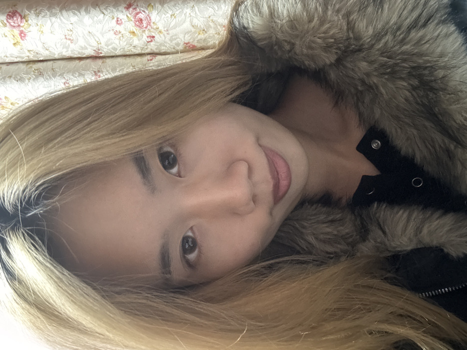 A close-up portrait features a female individual, likely of East Asian descent, looking directly into the camera. She has long, straight, blonde-colored hair with visible dark roots extending from the scalp. Her dark, almond-shaped eyes show reflections of a light source. She exhibits a subtle, closed-lip smile. The individual is dressed in a black garment, possibly a jacket, which has a prominent, light brown and grey faux fur collar or trim. Silver-colored snap fasteners are discernible on the black fabric beneath the fur. The background consists of a light-toned fabric curtain adorned with a repeating floral pattern of pink and red roses interspersed with green leaves, indicative of an indoor setting. The specific location within Shapingba District, China, cannot be determined from the visual information present in the image.