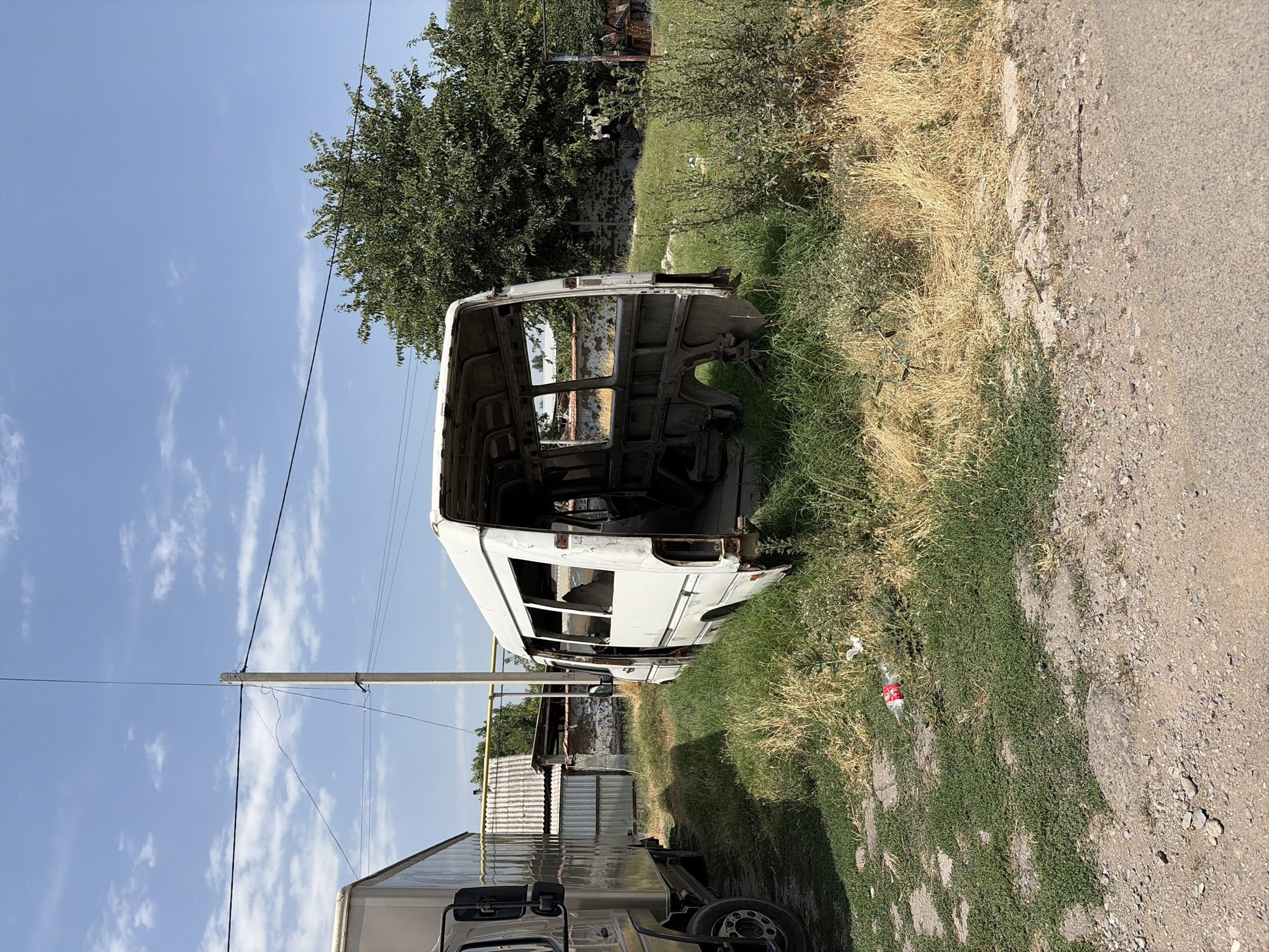 In Novopokrovka, Kyrgyzstan, an abandoned white minibus chassis sits in a grassy, overgrown area next to a parked truck. The minibus, stripped of its body panels and windows, reveals its internal frame and structure. The surrounding environment is characterized by dry grass, weeds, and scattered debris, including a plastic bottle near the minibus. A utility pole with overhead wires stands prominently in the background, behind the minibus, with a large green tree to the right. The sky is blue with scattered white clouds. The scene appears to be an outdoor junkyard or storage area for derelict vehicles.