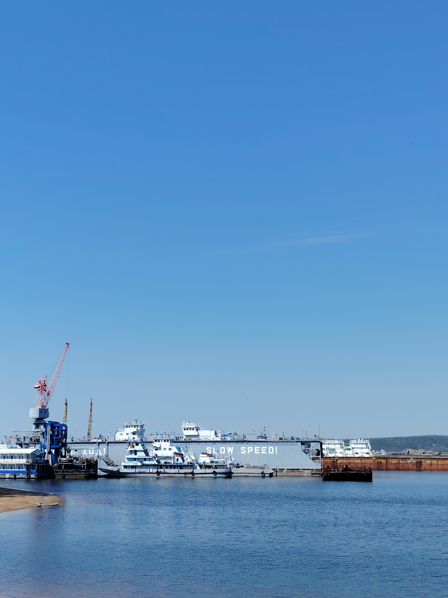 This photo shows a shipyard in the city of Chaikovsky, Russia. The scene features several ships moored at a dock, and a large crane positioned to the left of the vessels. The water is calm, and the sky is clear and blue.