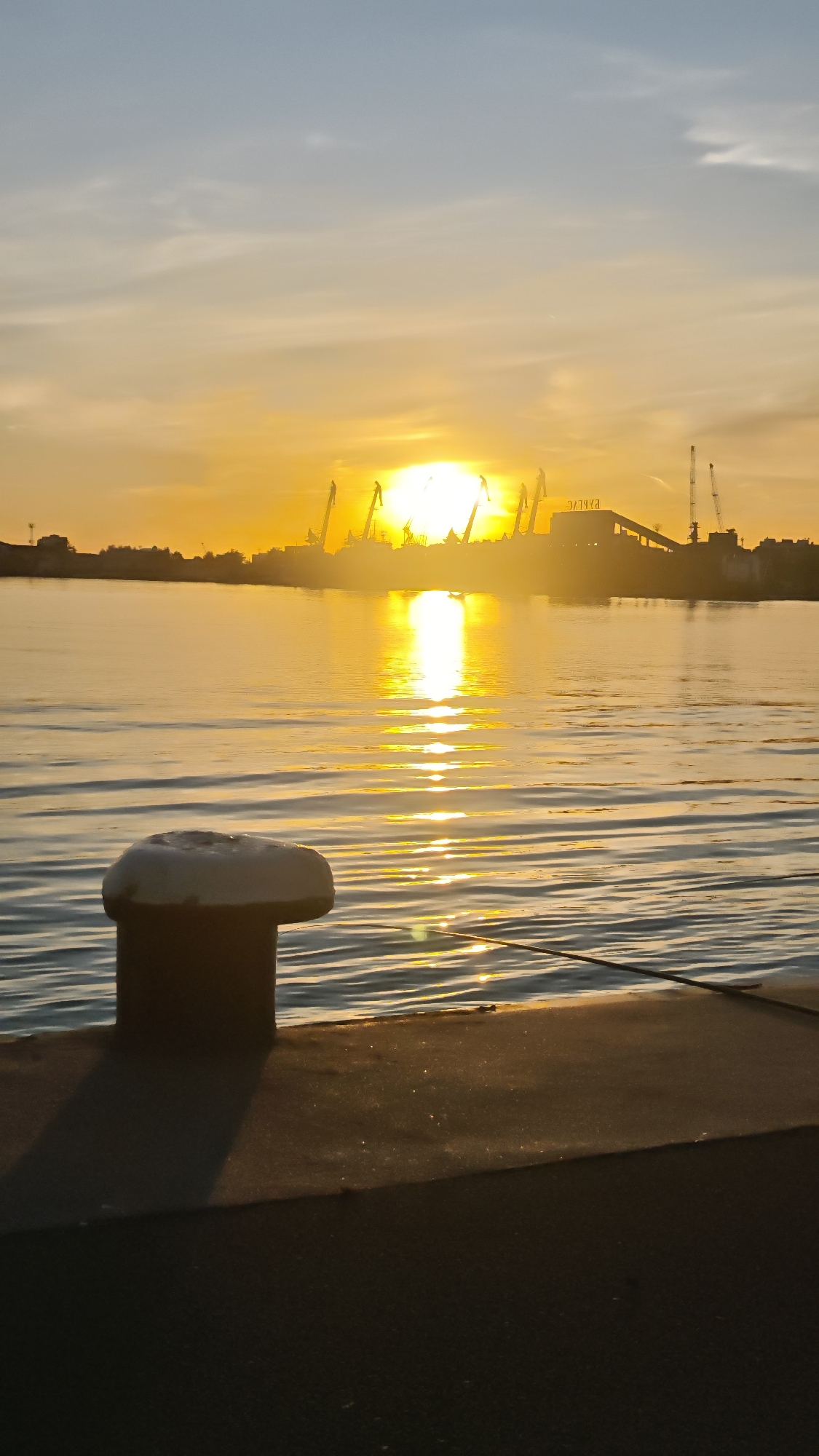 The image shows a sunset over a body of water, likely a bay or a harbor, in Burgas, Bulgaria. In the foreground is a dark, low pier with a prominent bollard used for mooring boats. The water surface has gentle ripples that reflect the golden light of the setting sun. In the background, a city skyline is visible, obscured by the sun's glare. Silhouettes of cranes and industrial structures suggest a port or industrial area. The sky is a gradient of blues and oranges, typical of a sunset. There are no people visible in the image.