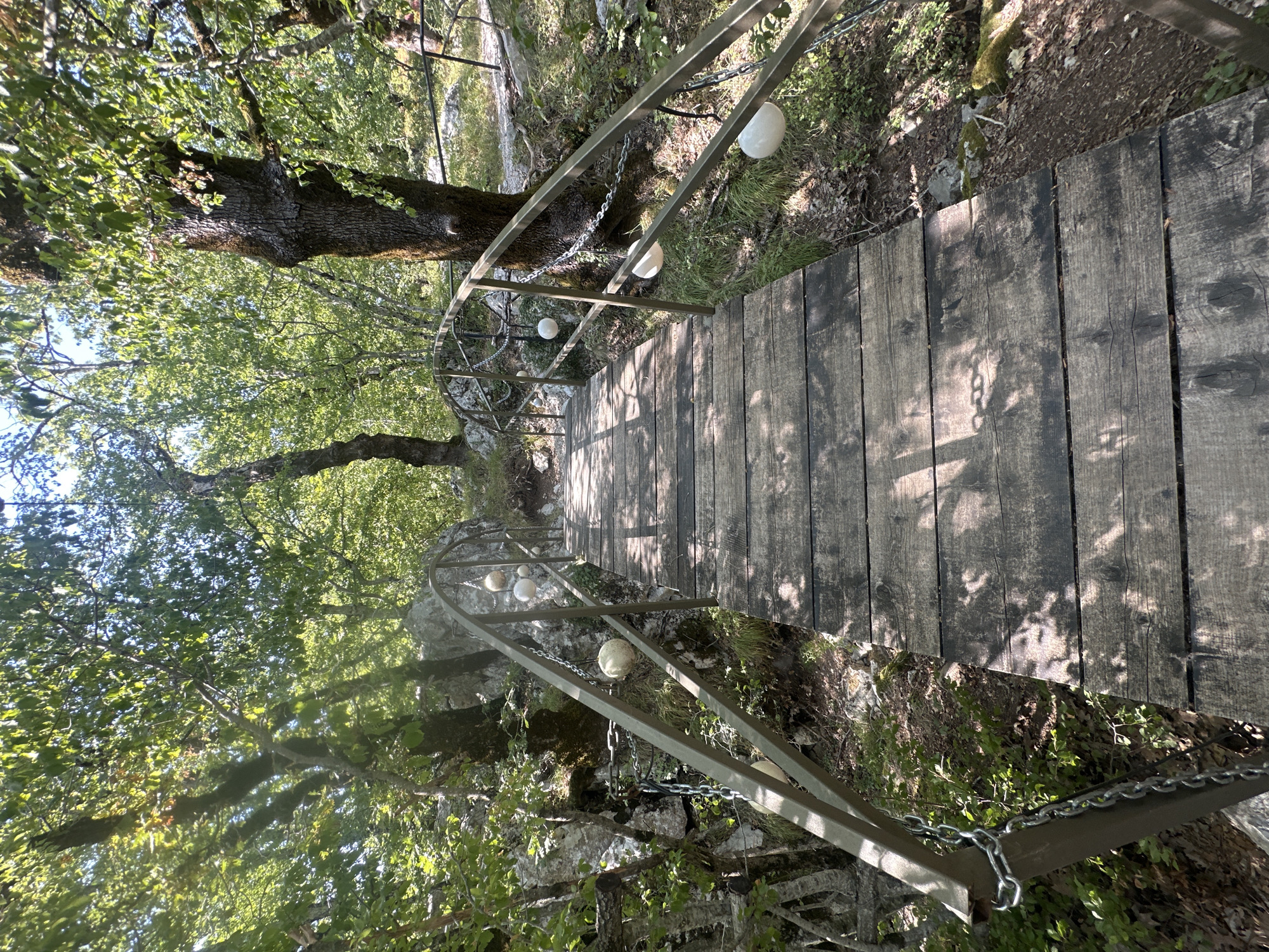 A wooden bridge with metal railings traverses a natural landscape, leading uphill and slightly to the right. The bridge's surface is constructed from weathered wooden planks, showing signs of wear and tear. Metal chains are used as decorative elements, hanging from the railings and supporting spherical white objects. The bridge is surrounded by lush green trees and vegetation, with sunlight filtering through the leaves, casting dappled shadows on the bridge. In the background, rocky outcrops and a path are visible, suggesting a hiking trail or a natural park setting. The overall scene depicts an outdoor environment with a man-made structure integrated into the natural surroundings. The location is identified as being in the city of Bukovik, Montenegro.