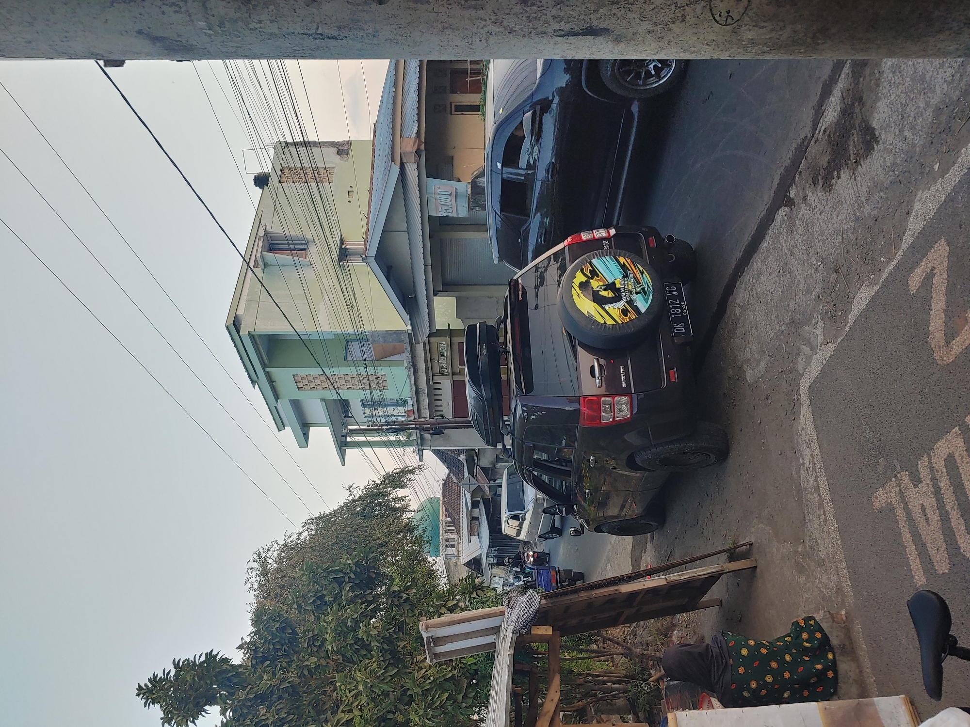 A street scene in Kembang Kerang, Indonesia, is depicted with traffic and buildings. In the foreground, a person wearing a dark headscarf and a green floral-patterned dress sits on the ground next to a wooden structure. A dark-colored Suzuki SUV, with a spare tire cover featuring a yellow and blue graphic, is parked on the right side of the road. To its right, a dark SUV is also visible. Further down the road, a white SUV is in motion, followed by a blue cart with passengers and a motorcycle. A building with a green facade and multiple windows is prominent in the background, with a green-domed building, possibly a mosque, further in the distance. Overhead, power lines crisscross the sky, and trees line the left side of the street. The ground is a mixture of dirt and asphalt.