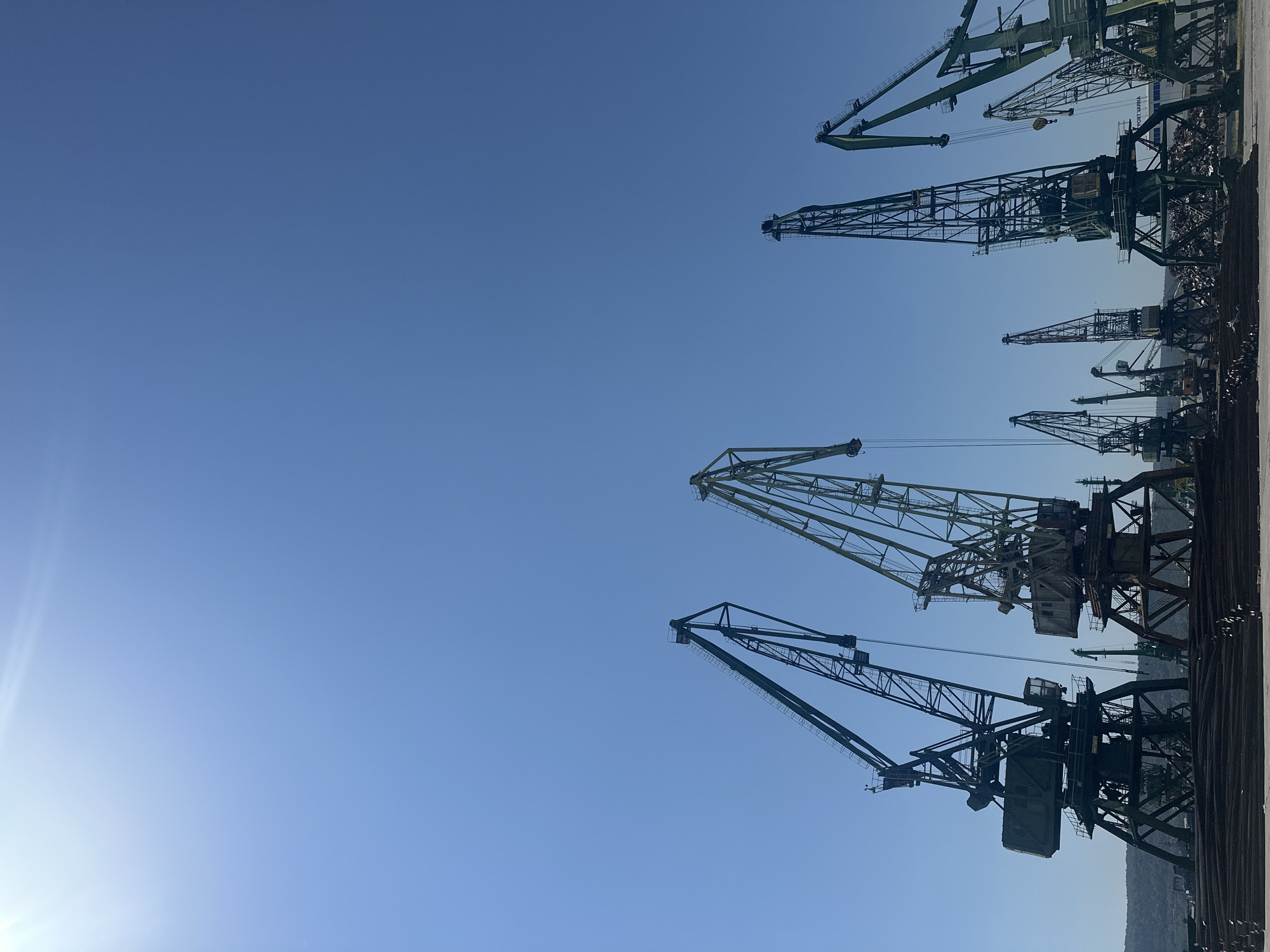 The image shows a port with several large cranes silhouetted against a clear blue sky. The cranes are of various sizes and designs, with some appearing older and more weathered than others. In the foreground, a large stack of metal rebar or similar material is visible. The background shows a shoreline with hills or mountains. The sun is shining brightly, casting a slight lens flare in the upper left corner of the image. The scene suggests an industrial area within a port.