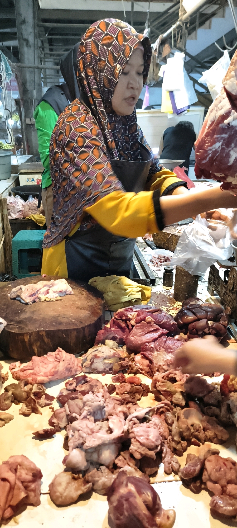 The image depicts a butcher stall in the Weru Lor market, Indonesia. A woman wearing a hijab with a geometric pattern of orange, purple, and white, and a yellow shirt with black trim, stands behind a counter laden with raw meat. She wears a dark apron. To her left, a large, round wooden chopping block is visible, with a piece of pale meat on top. The counter displays various cuts of raw red meat and offal, including what appears to be liver and intestines. In the background, another person in a green shirt and a person in a black shirt are partially visible. Bags and other market items are also present in the background. The lighting suggests an indoor market setting, possibly with overhead fluorescent lights. The overall scene is of a busy food market.
