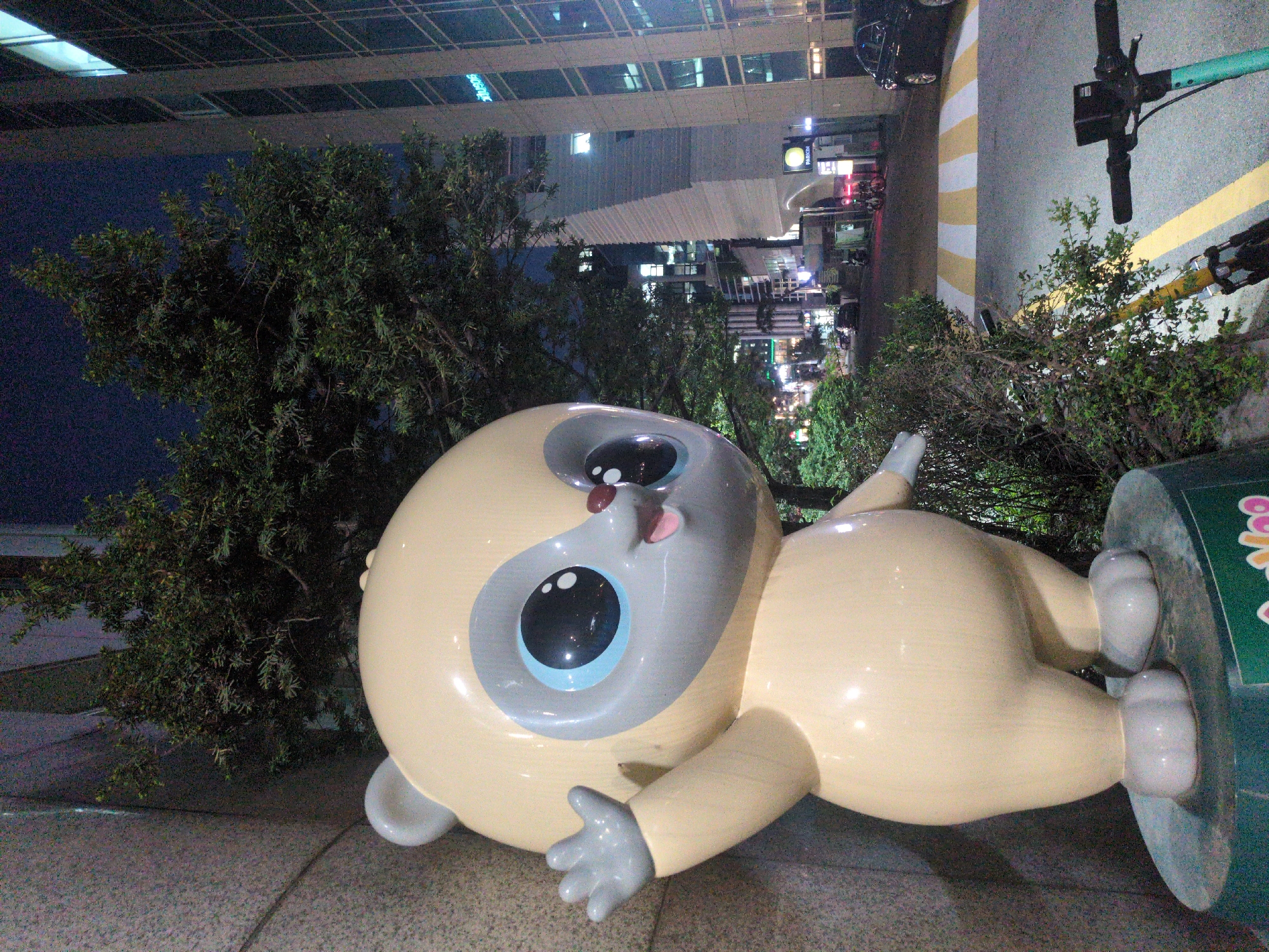 The image depicts a whimsical statue of a cartoonish character in Seoul, South Korea. The statue is lying on its stomach with a large, round head, big eyes, and a small, protruding tongue. It has pale yellow skin and gray features around its eyes, hands, and feet. The statue is positioned on a gray tiled surface, with a tree to its left and bushes behind it. In the background, there are city buildings with illuminated windows, and a street with crosswalk markings and electric scooters. The overall lighting suggests it is night time.