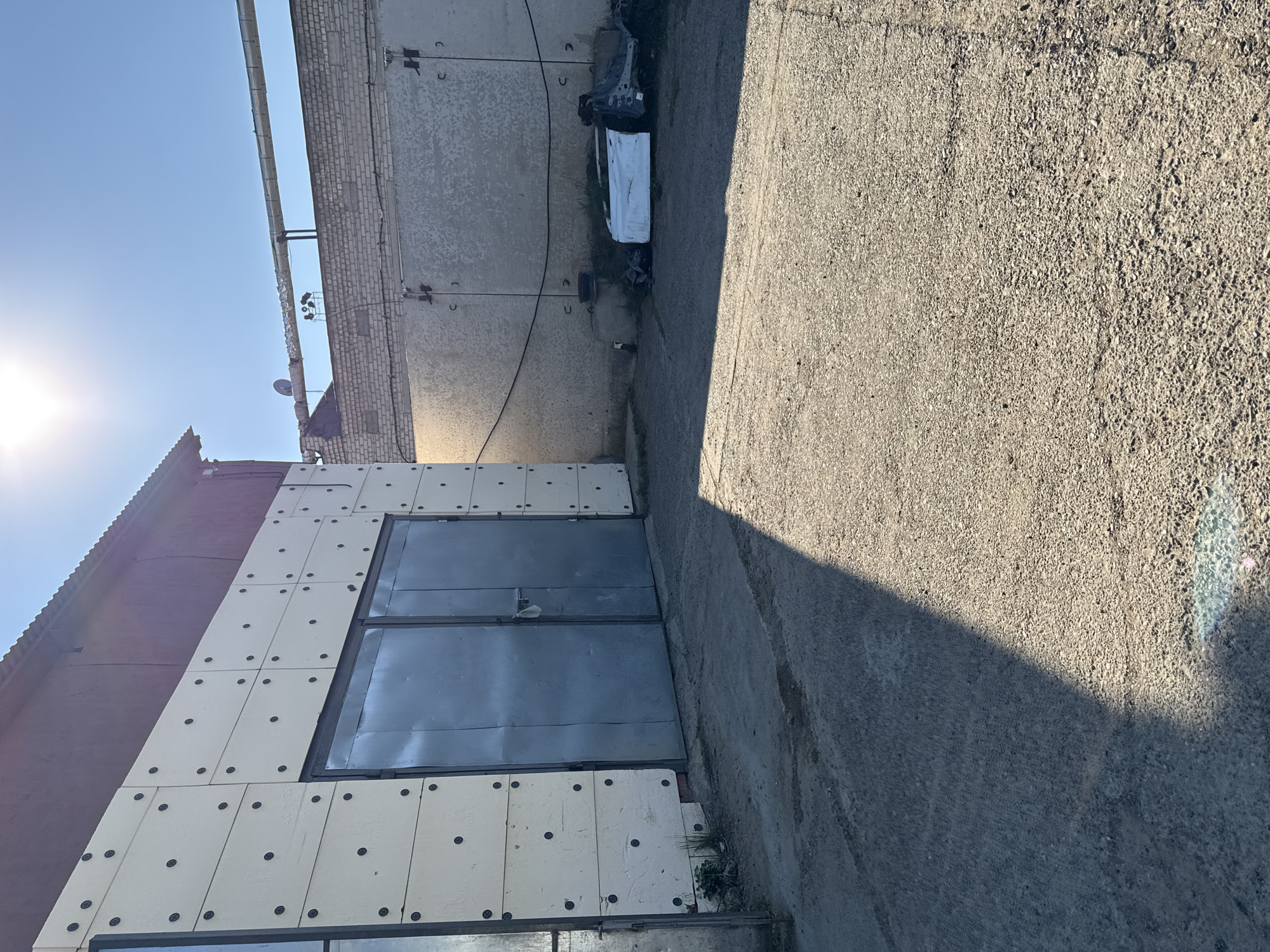 The image shows an industrial area in Chaikovsky, Russia, featuring a concrete or asphalt driveway adjacent to a building. The building's facade consists of a dark red upper section and a lower section clad in white panels with dark circular fasteners. A large, dark gray metal door is prominent on the building's side. The adjacent wall is a plain concrete structure with various wires and metal attachments. In the space between the building and the wall, some discarded car parts are visible. The scene is illuminated by sunlight, creating a distinct shadow across the driveway. The sky is clear and blue.