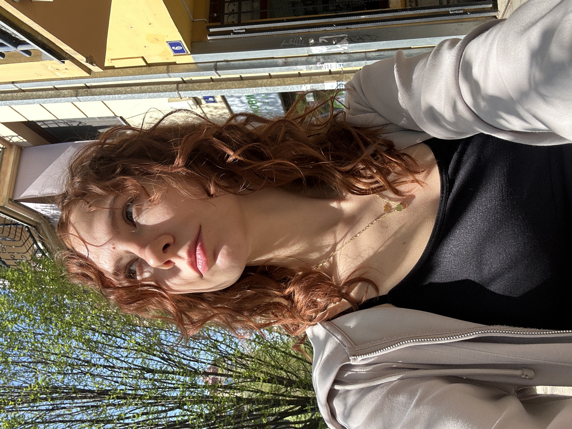 A young woman with reddish-brown, curly hair is posing for a photo outdoors on a sunny day. She is wearing a black top and a light grey jacket. She has a gold necklace with small pendants. In the background, blurred due to the depth of field, there are green trees and a building. The building has a visible address plaque with the number "9" on it, indicating the location is in Novi Sad, Serbia.